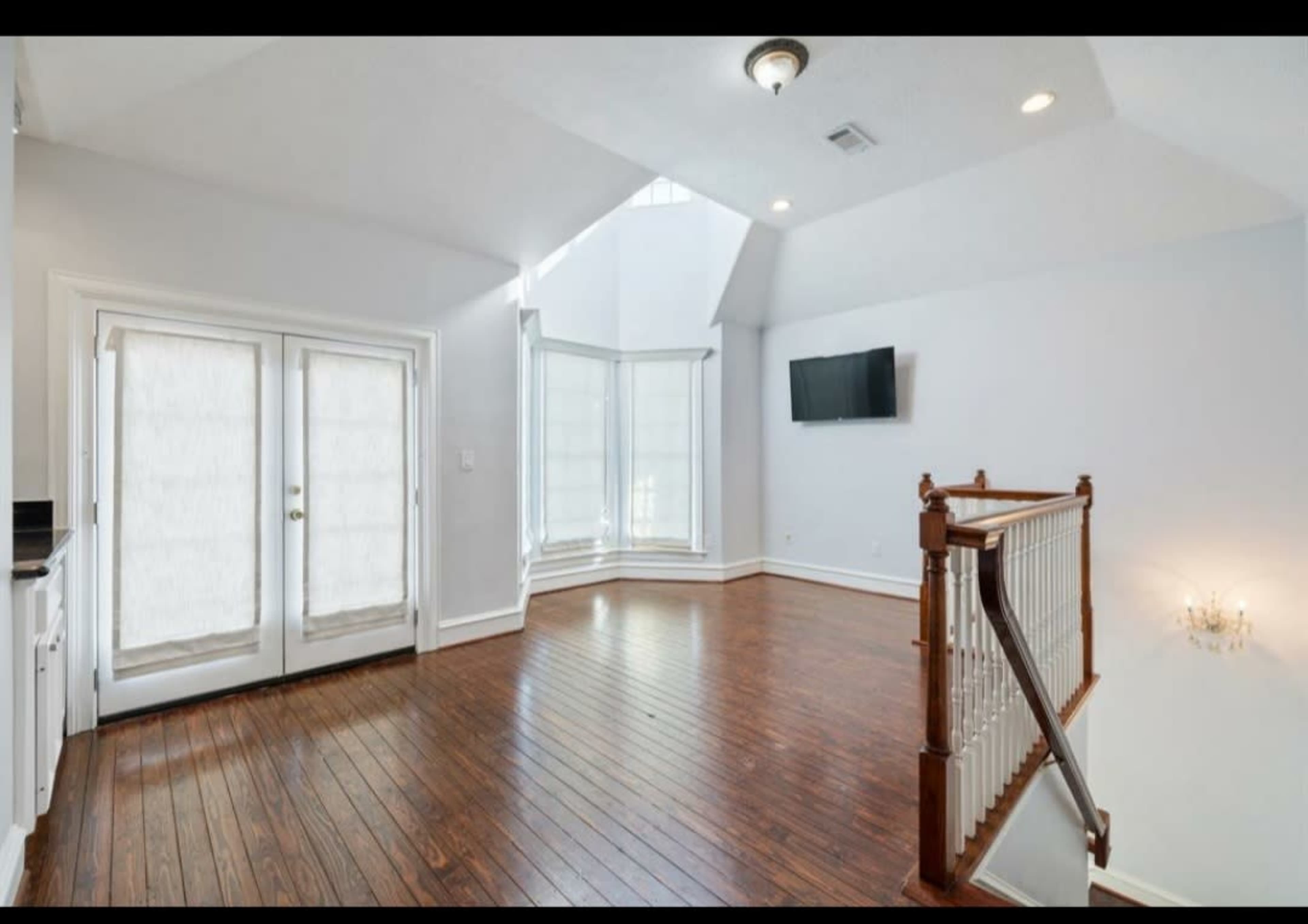 A spacious, empty room with hardwood floors, large windows, and a television mounted on the wall.