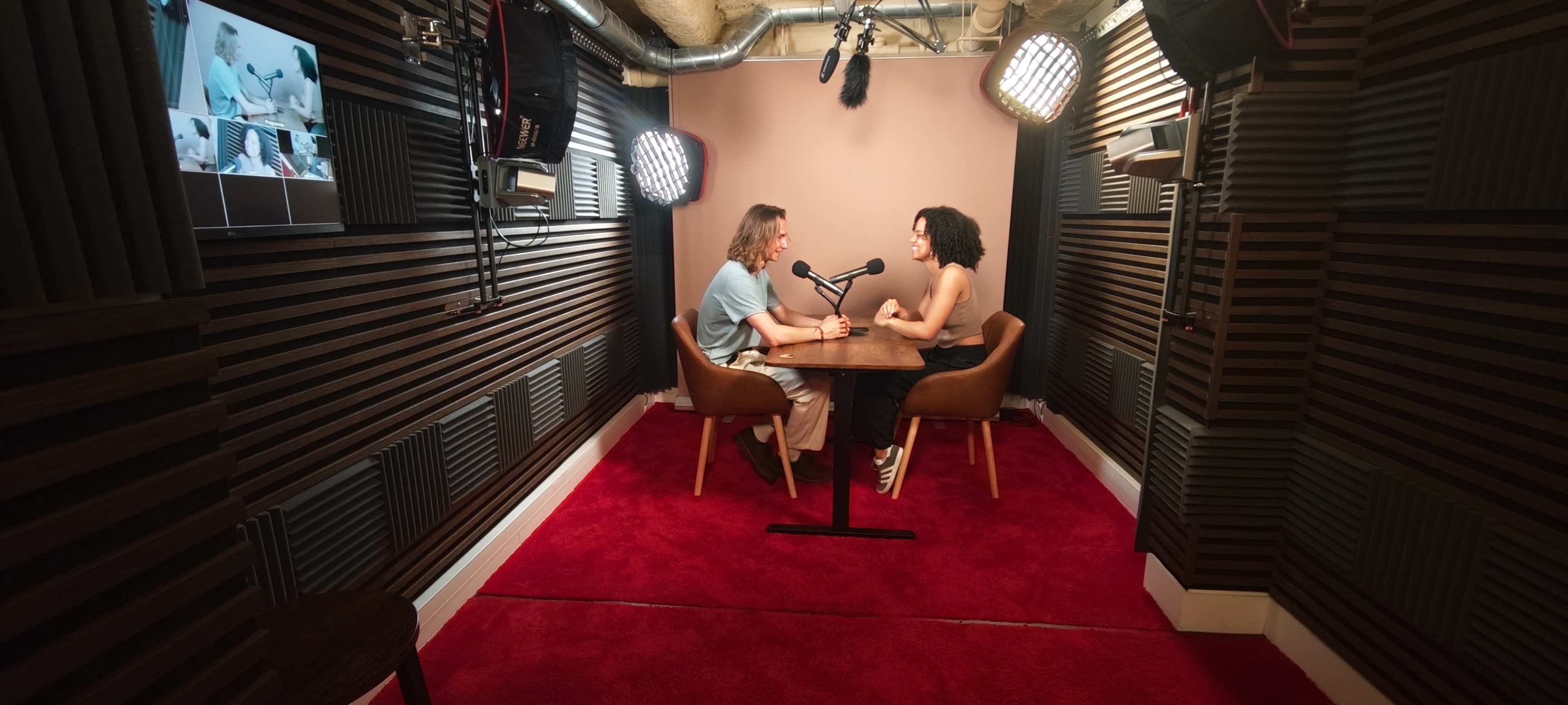Two people are seated at a table in a soundproof studio, facing each other under bright lights.