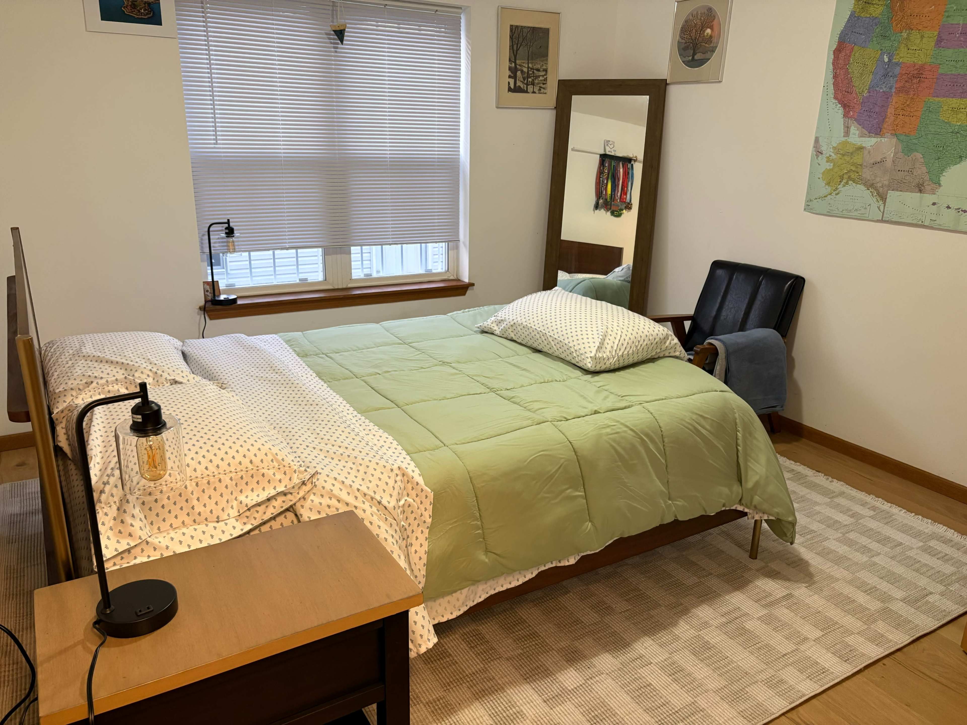 The image shows a bedroom with a neatly made bed featuring a green comforter, a mirror, a chair, and a window with blinds.