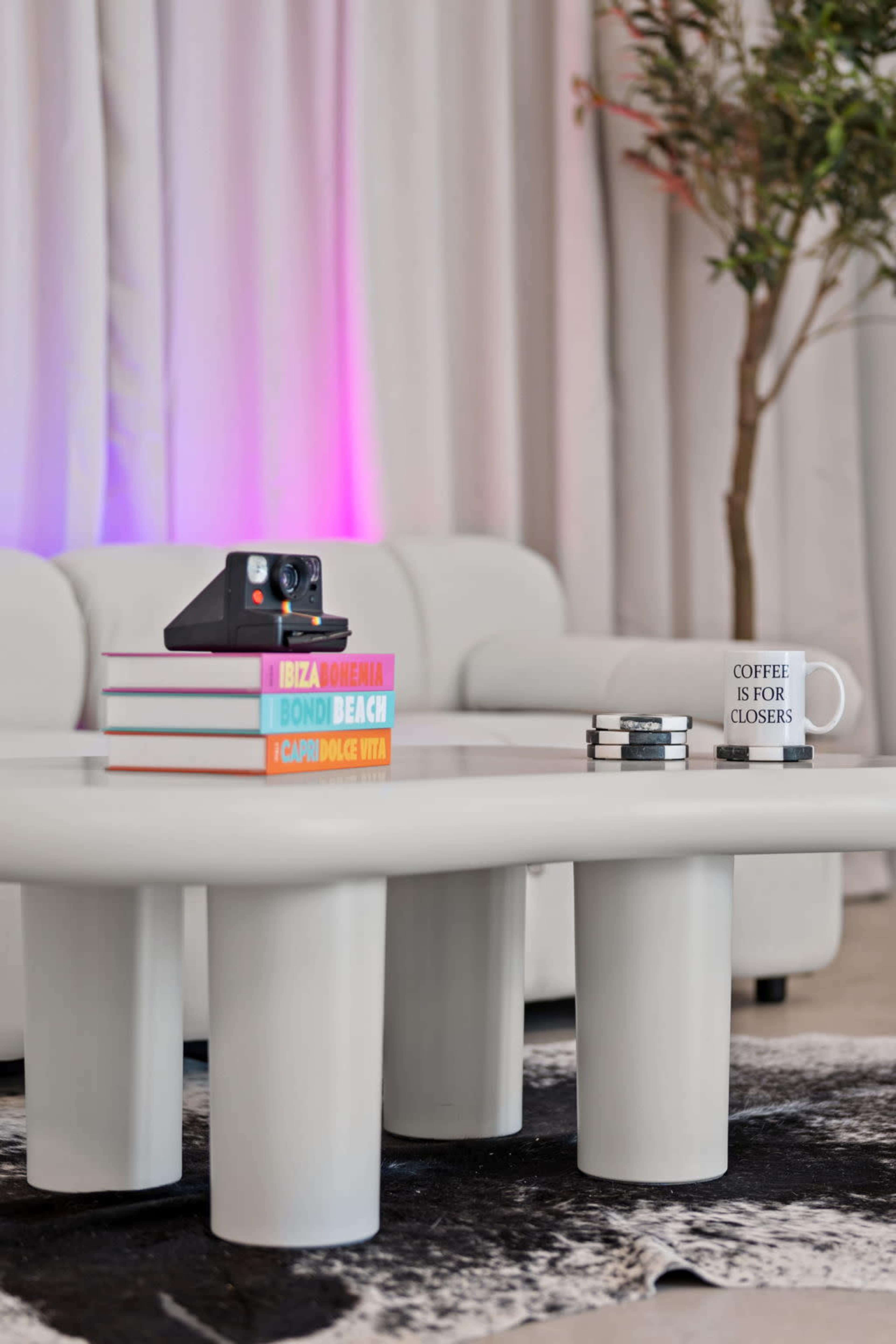 A modern coffee table holds stacked colorful books, a camera, coasters, and a mug that reads "COFFEE IS FOR CLOSERS."