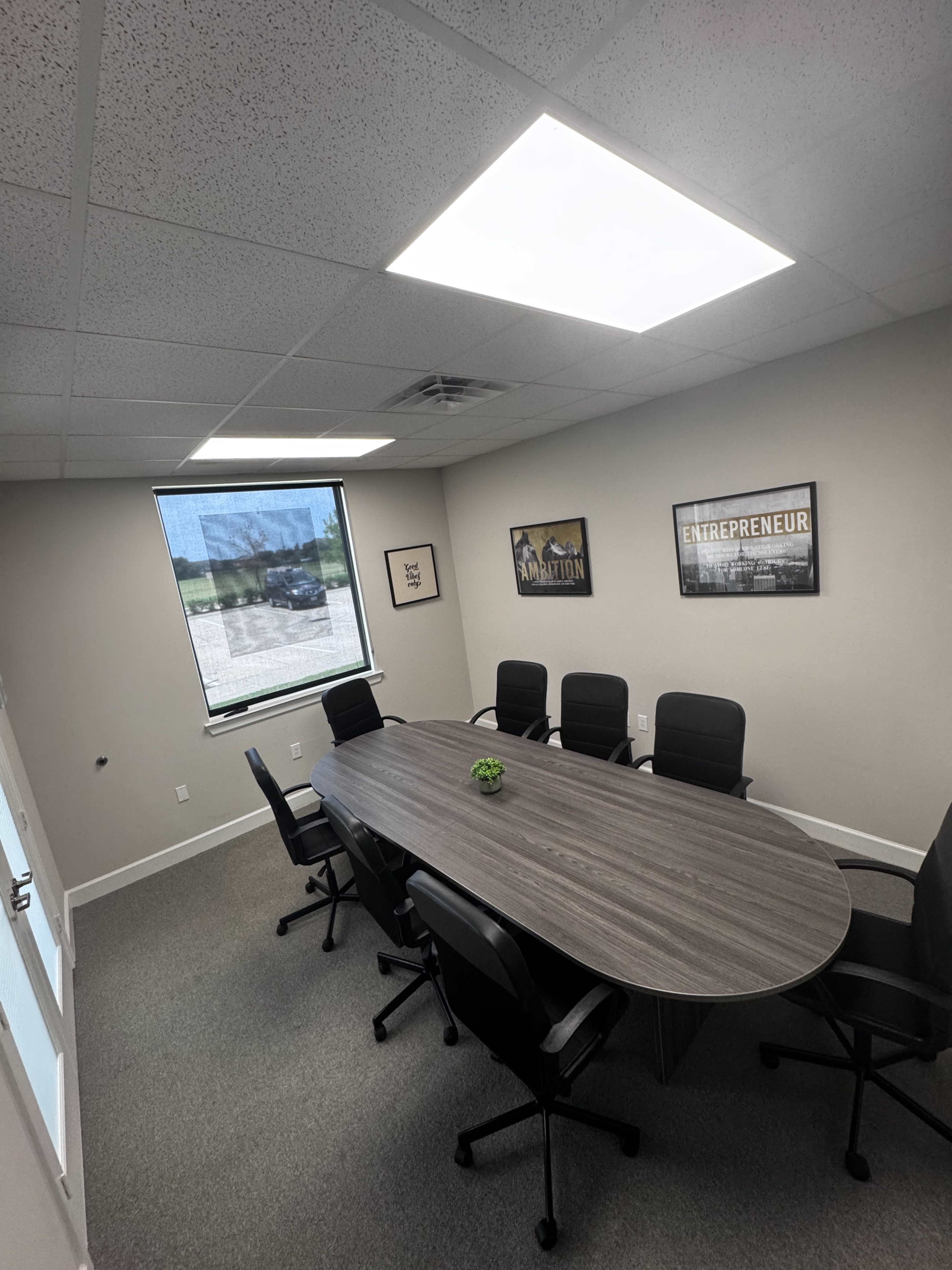 A conference room features a long, oval table surrounded by six black chairs, with a large window and two framed posters on the walls.