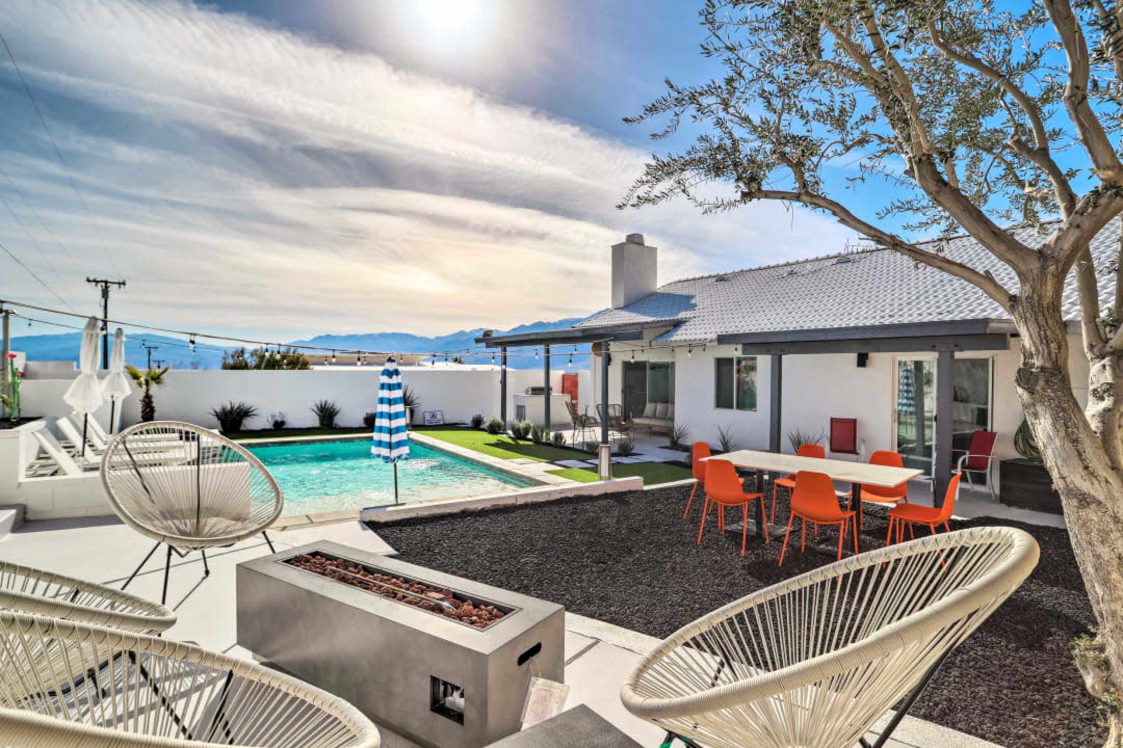 The image features a modern outdoor space with a swimming pool, patio furniture, and a landscaped area surrounded by mountains under a cloudy sky.