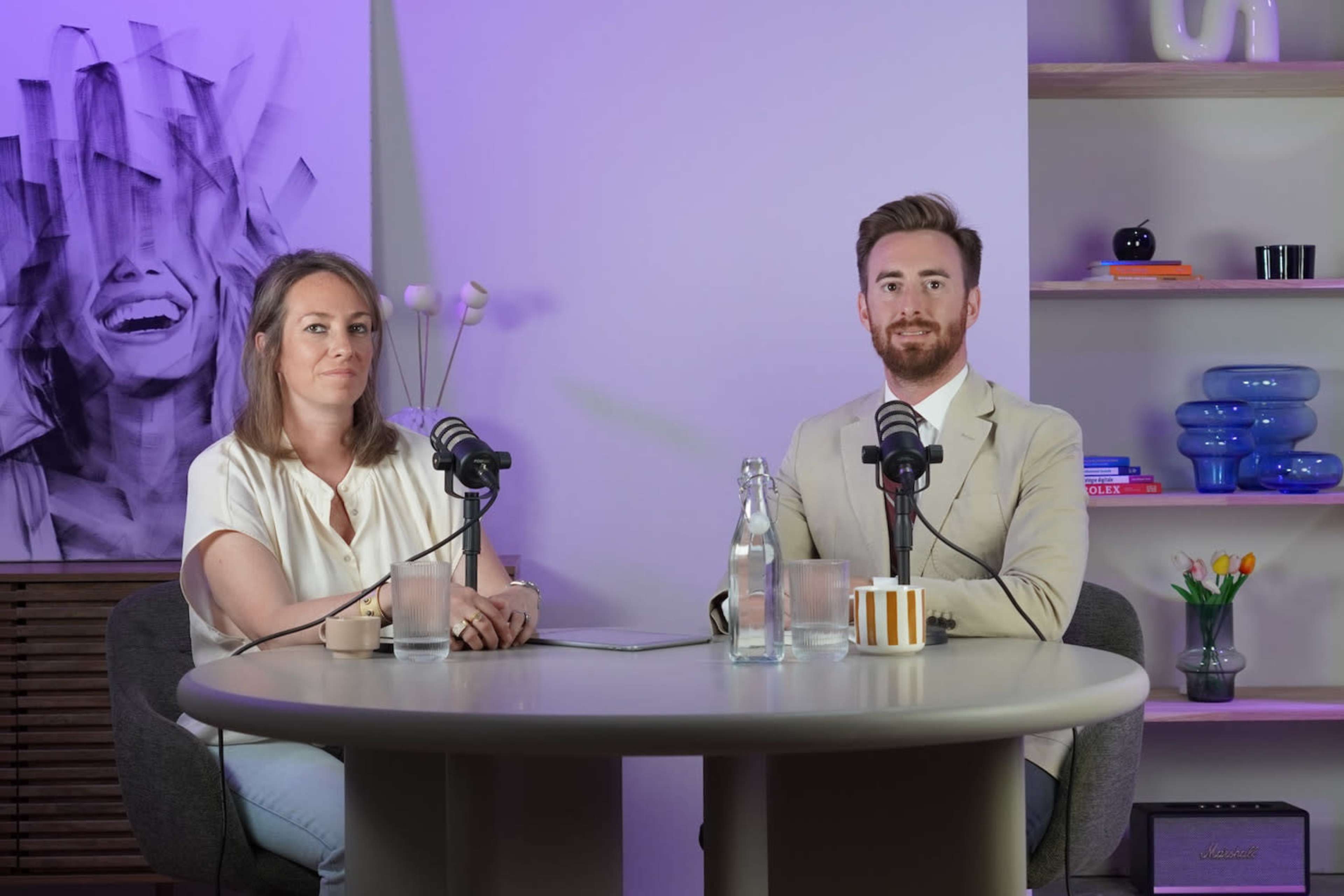 A woman and a man are seated at a round table with microphones in front of them, in a well-lit room featuring colorful decor and a large artwork on the wall.