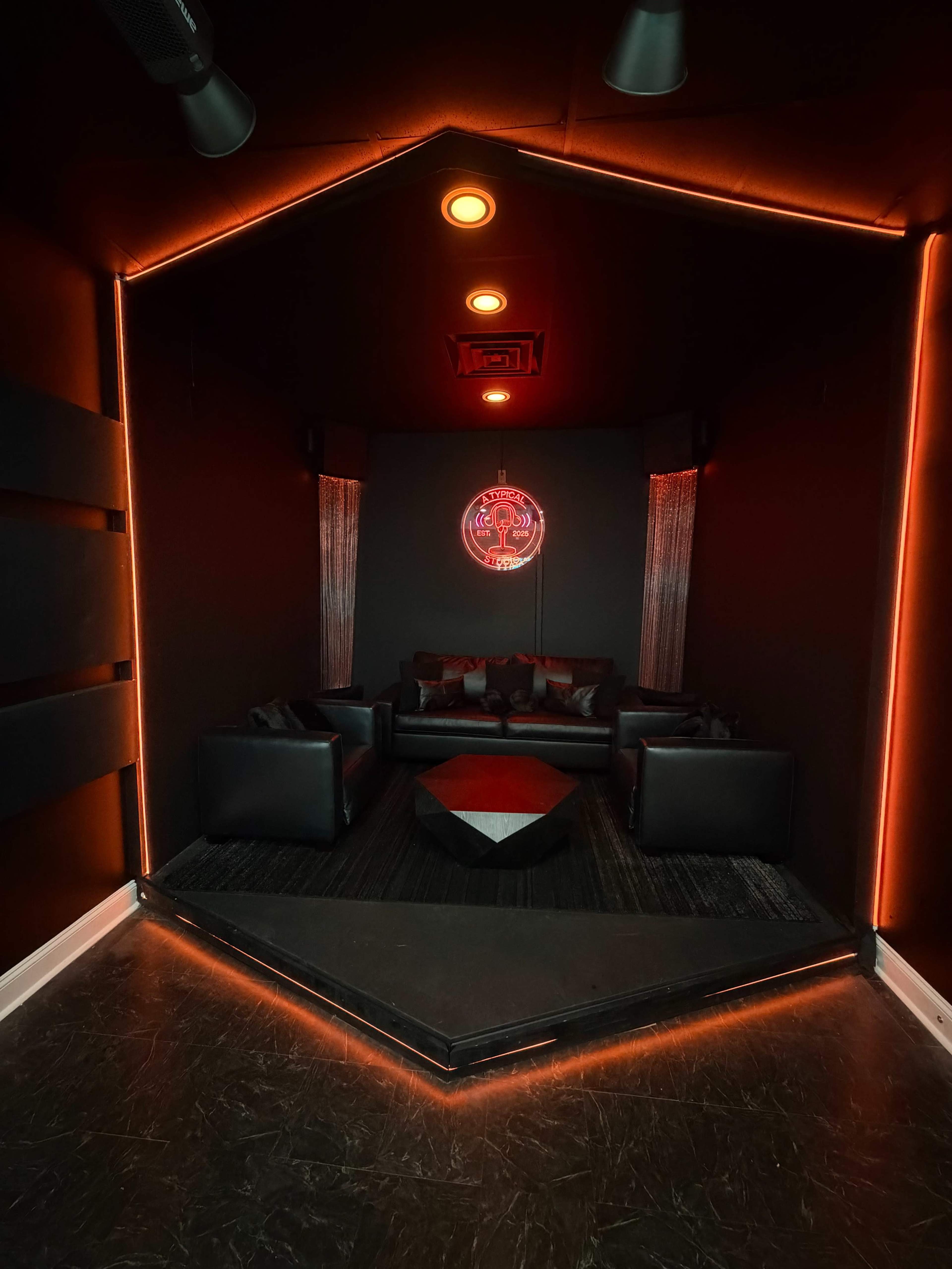 The image depicts a dimly lit lounge area with black walls, a striking neon sign, and modern furniture arranged around a central geometric table.