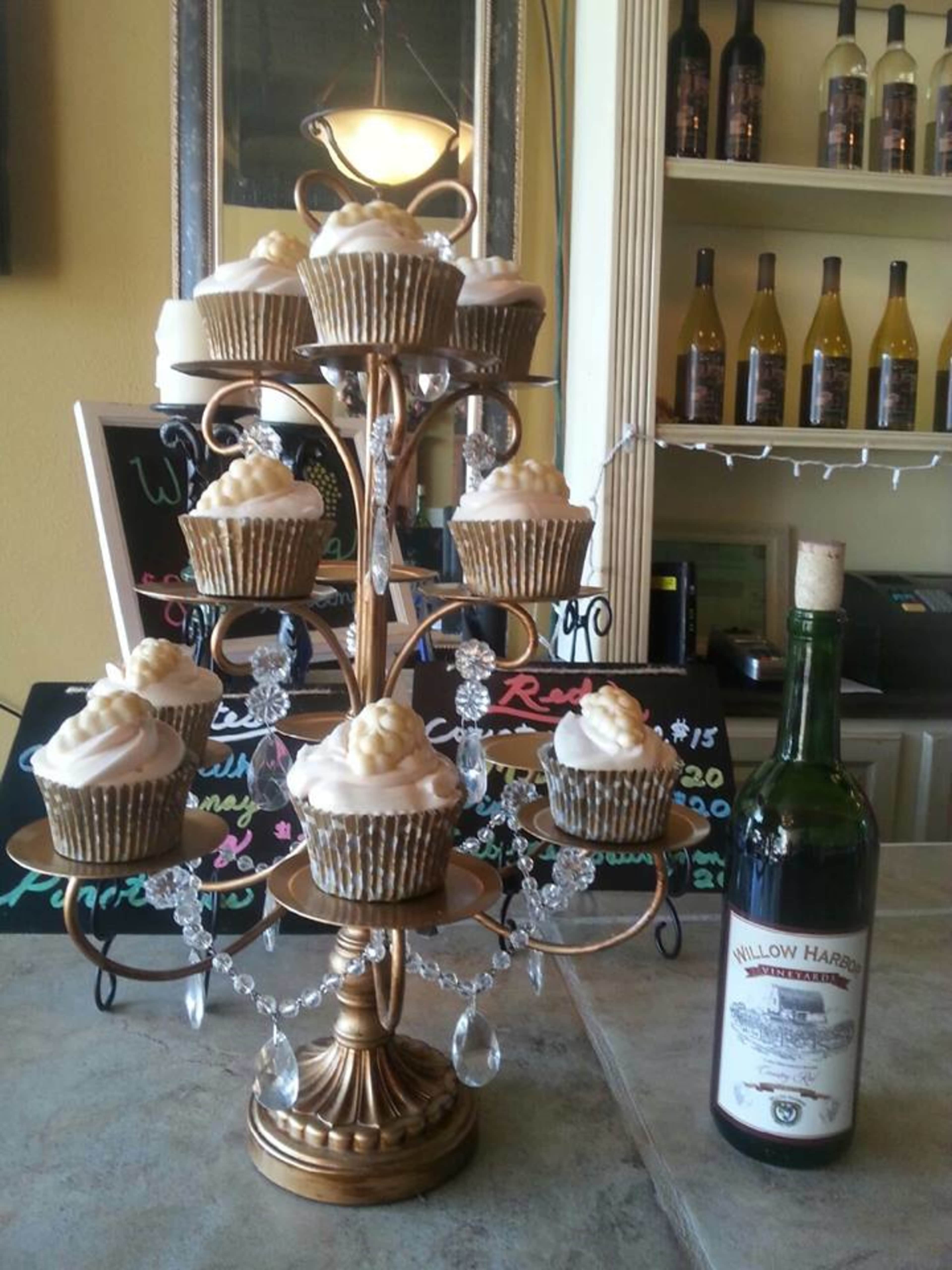 A decorative stand holds twelve cupcakes topped with frosting, surrounded by crystal accents, next to a bottle of wine on a countertop.