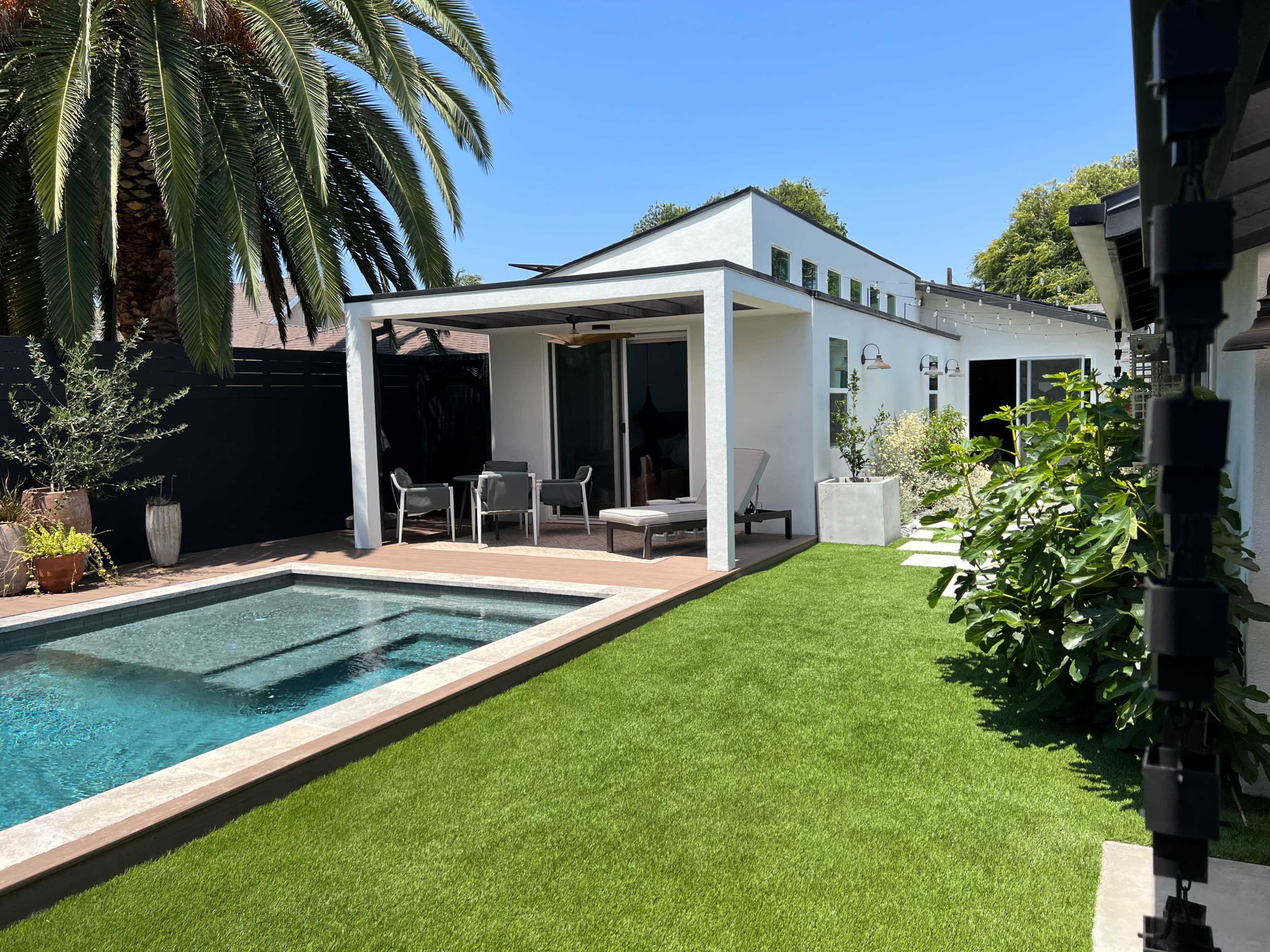 A modern backyard features a swimming pool, green grass, and a covered seating area adjacent to a white house.