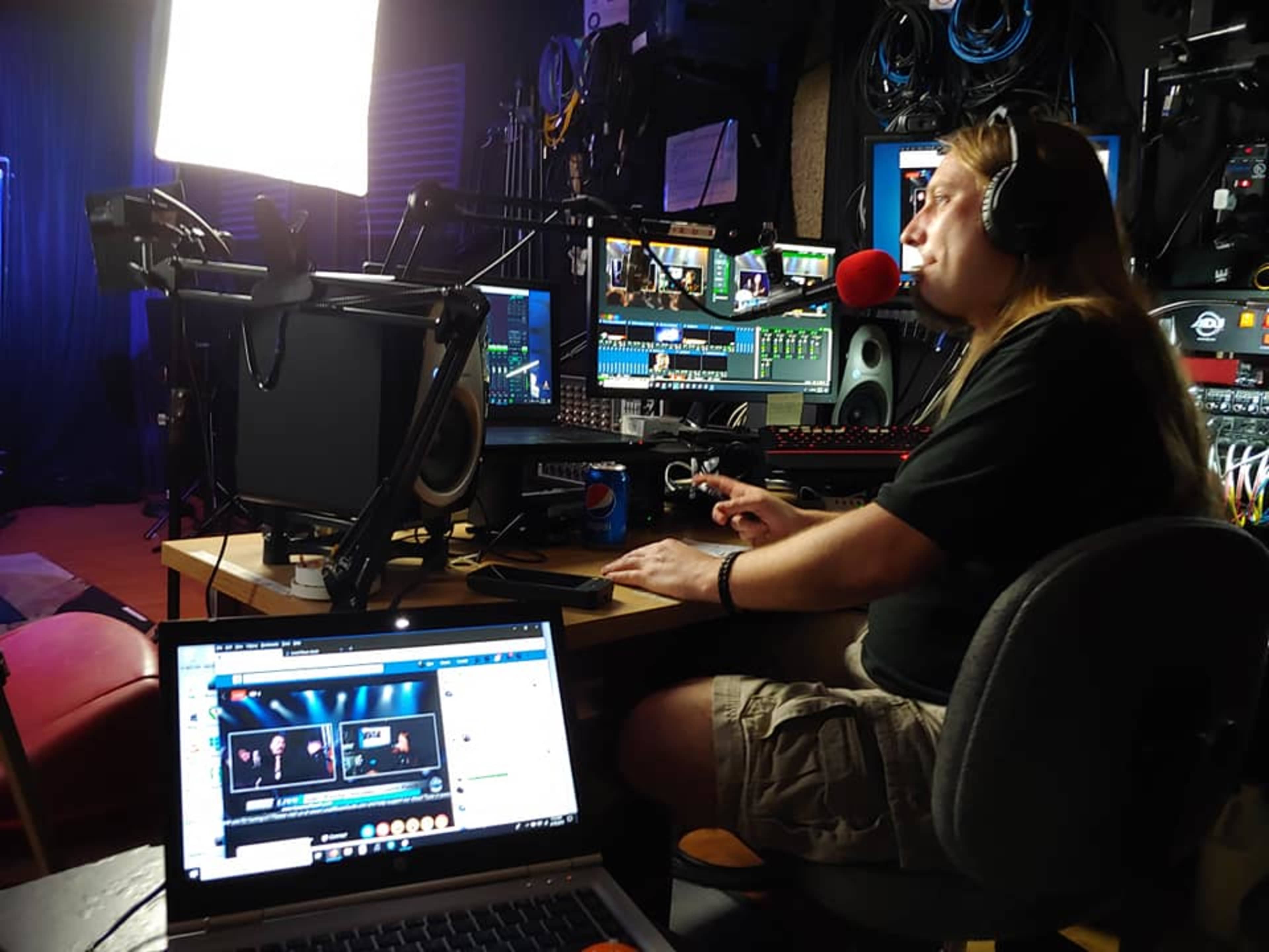 A person with long hair operates a video production setup featuring multiple screens and equipment in a dimly lit studio.