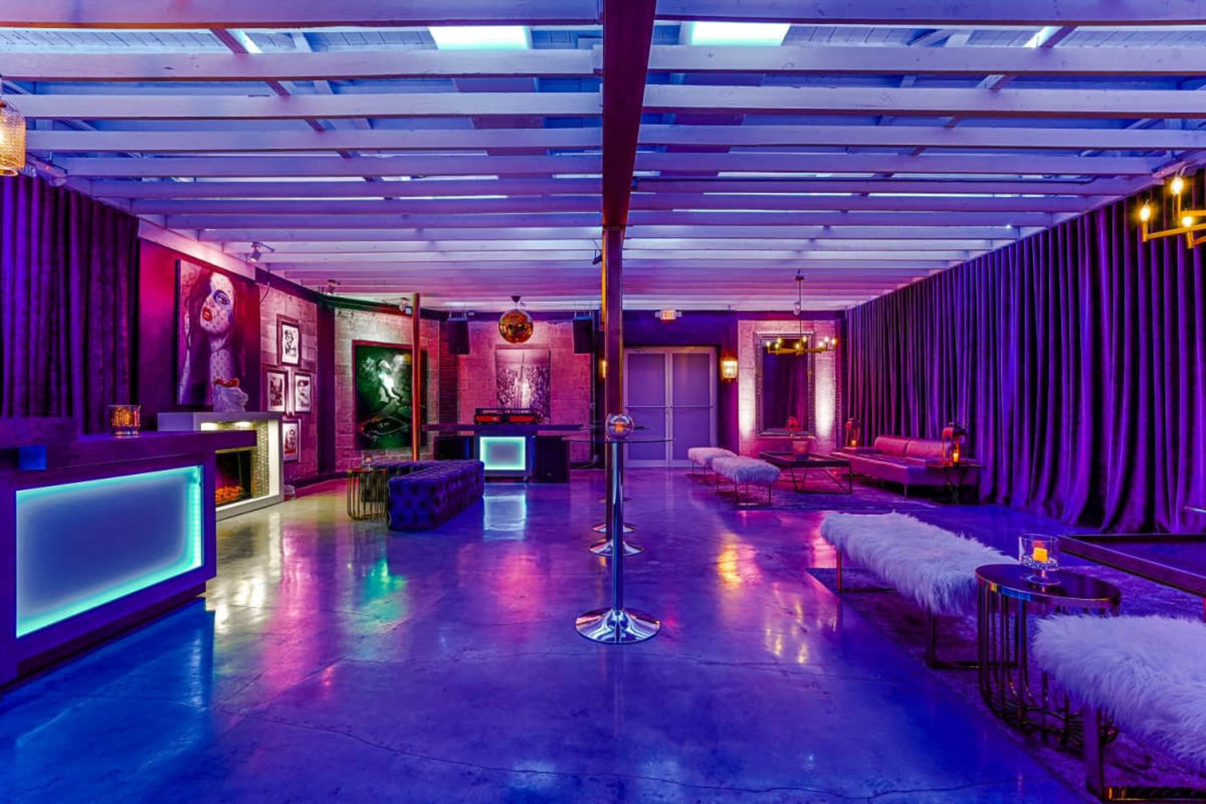 The image shows a dimly lit lounge space featuring purple lighting, plush seating arrangements, and artwork displayed on the walls.