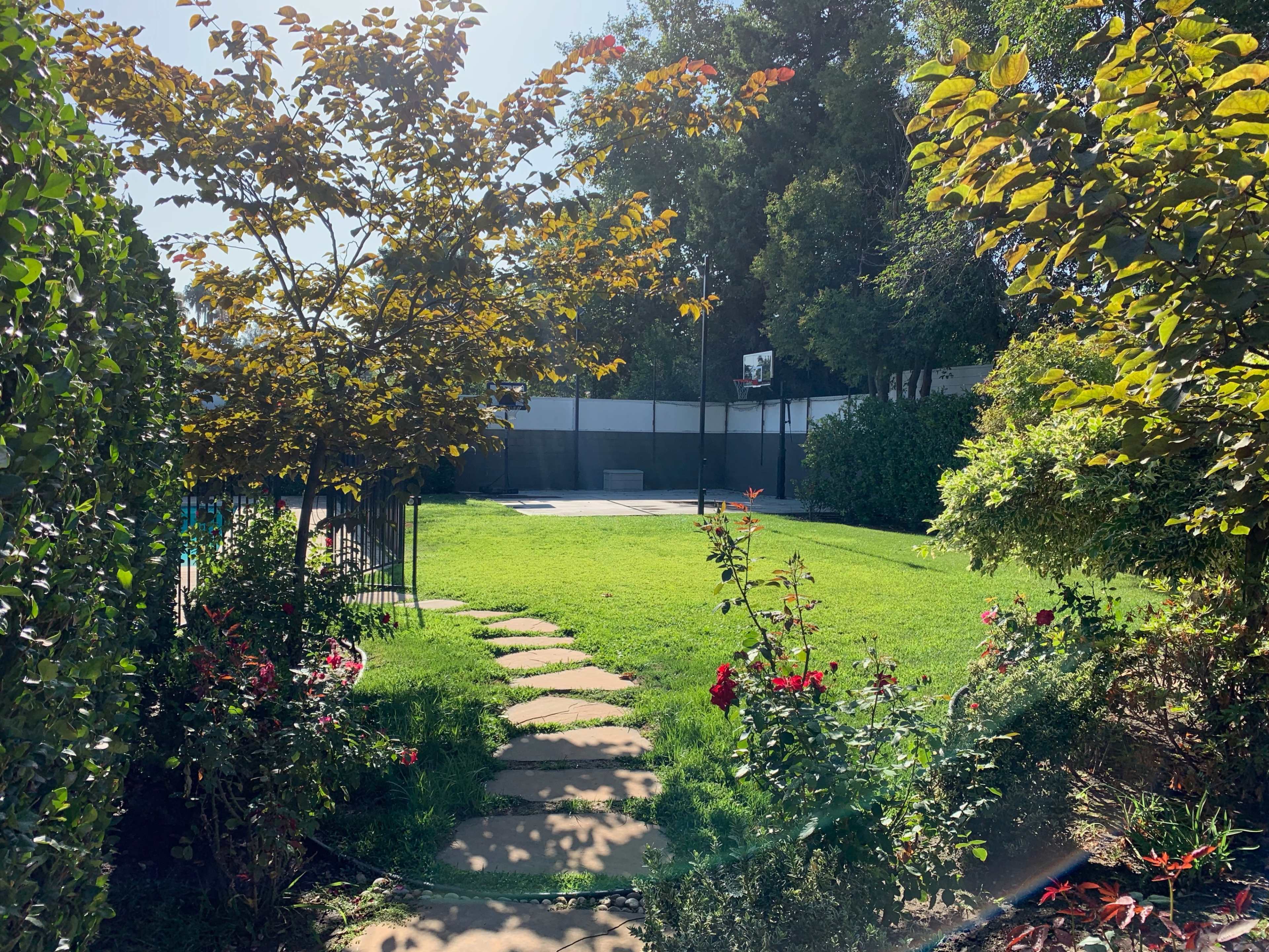 A backyard with a grassy area, a basketball hoop visible in the background, and flower beds along a stone pathway.