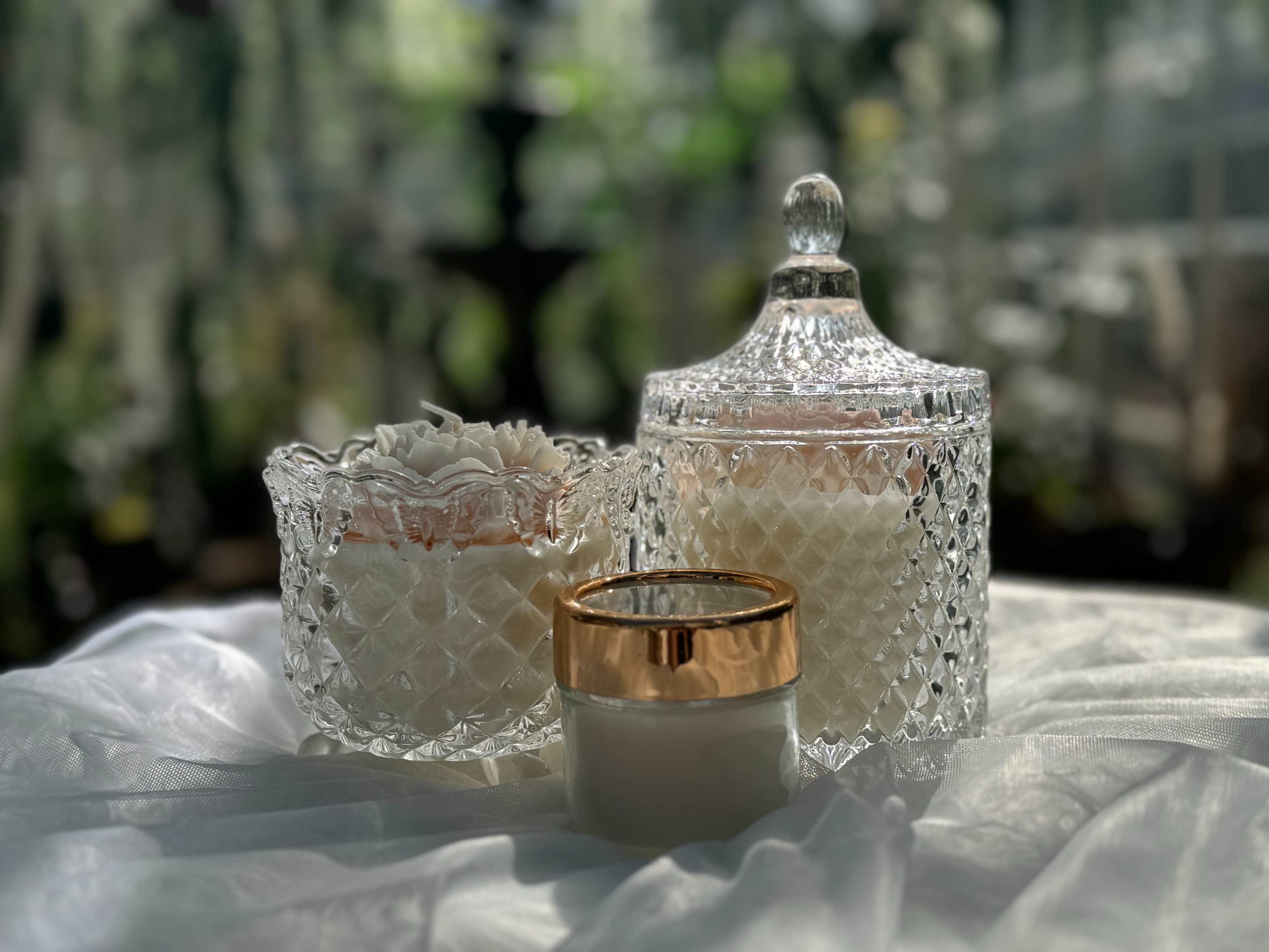 Two decorative glass containers, one with a lid and intricate designs, alongside a smaller jar with a gold band, all placed on a light fabric.