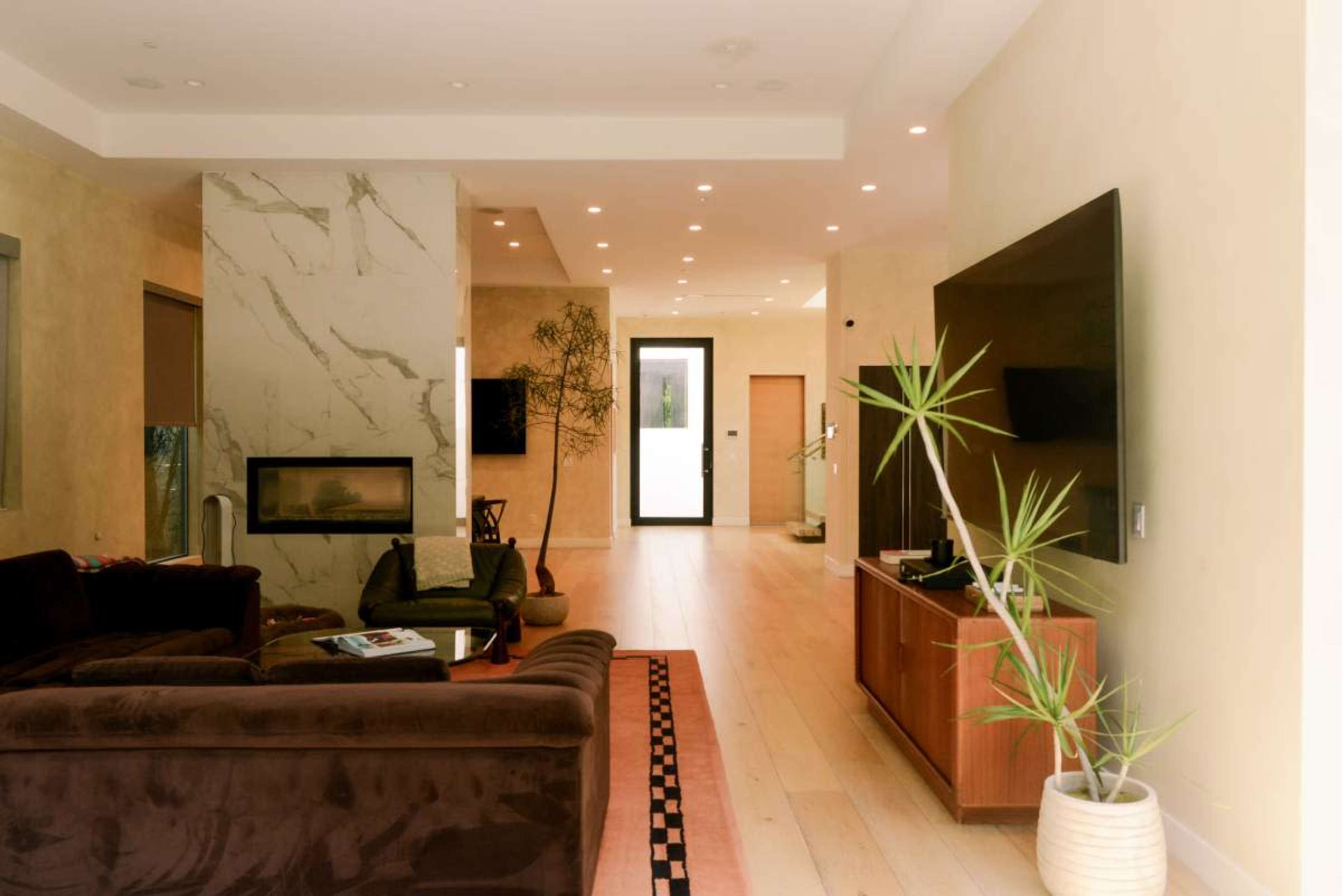 The image shows a modern, open-concept living room with a dark sofa, a mounted television, a minimalist coffee table, and large windows offering natural light.