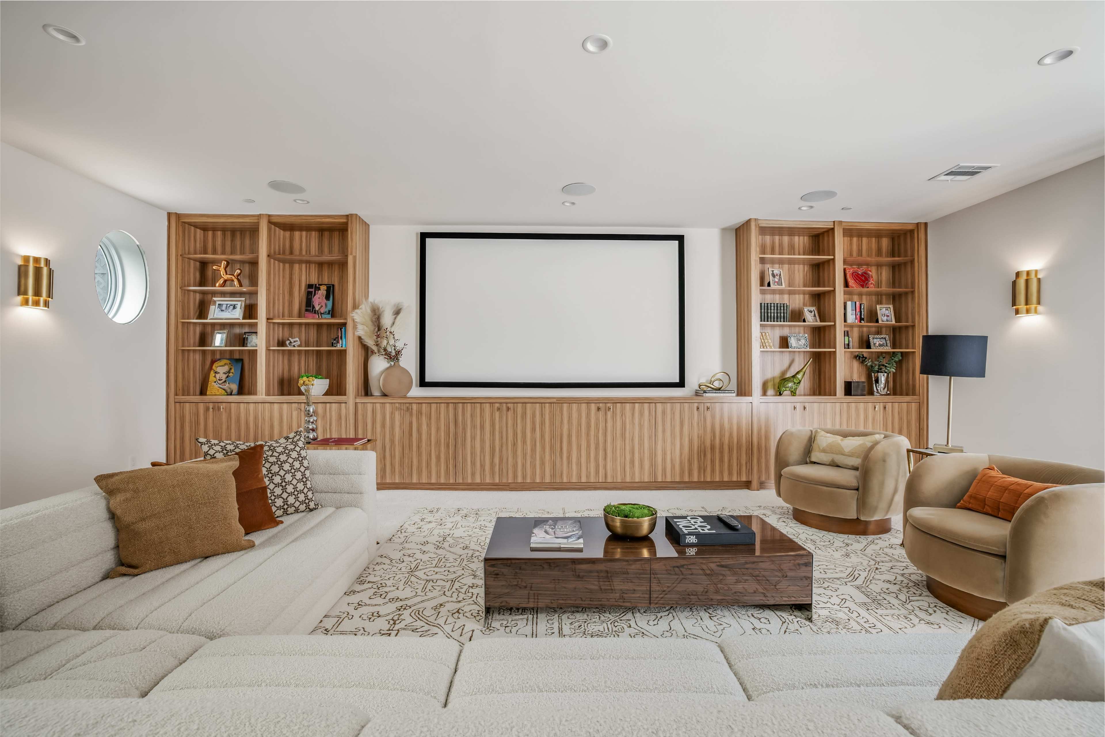 The image shows a modern home theater with a large wall-mounted screen, wooden bookshelves filled with books and decor, a coffee table, and cozy seating including a sectional sofa and two armchairs.