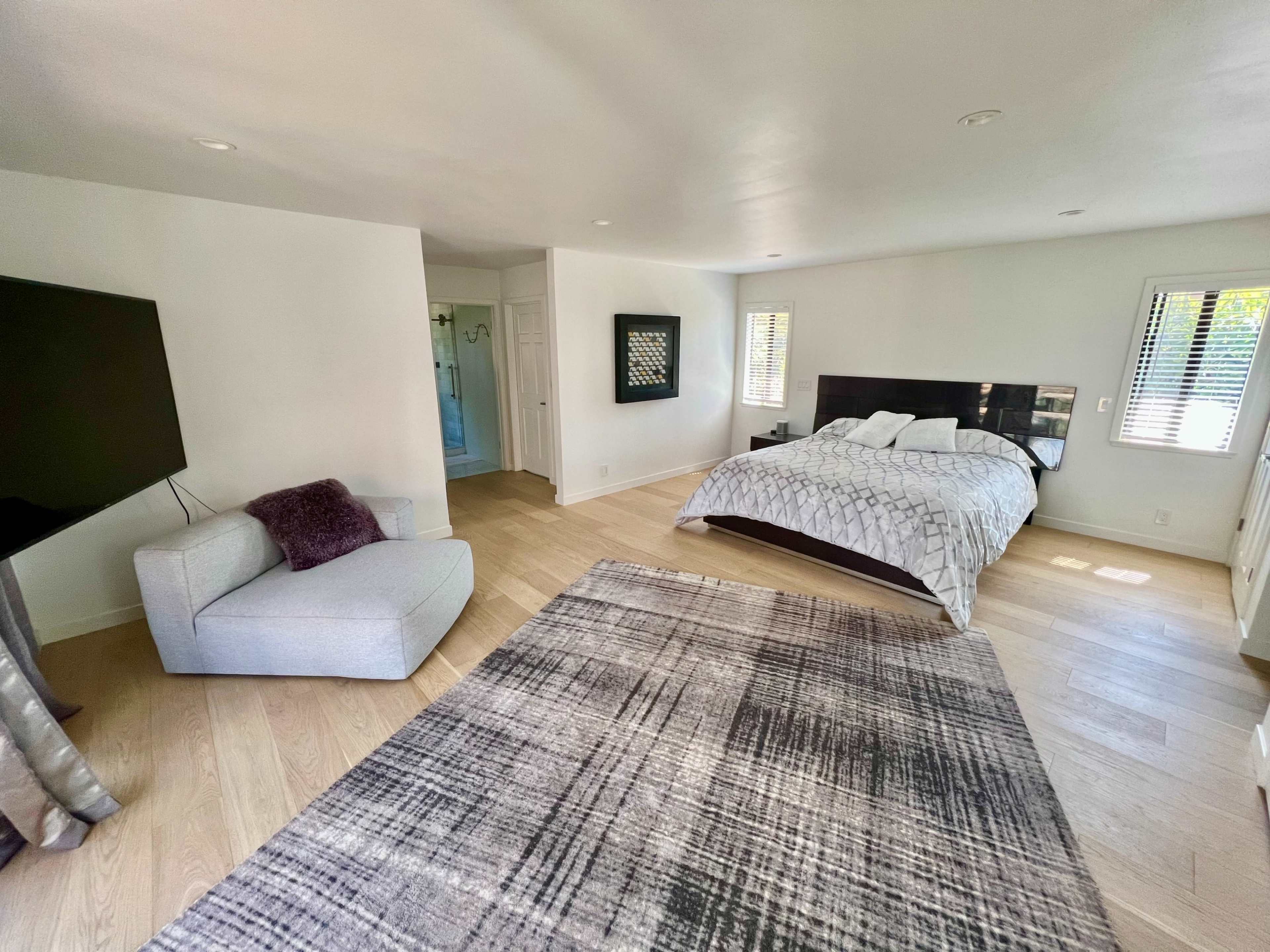 A spacious bedroom featuring a large bed, a flat-screen television, a light-colored armchair, and a patterned rug on the wooden floor.