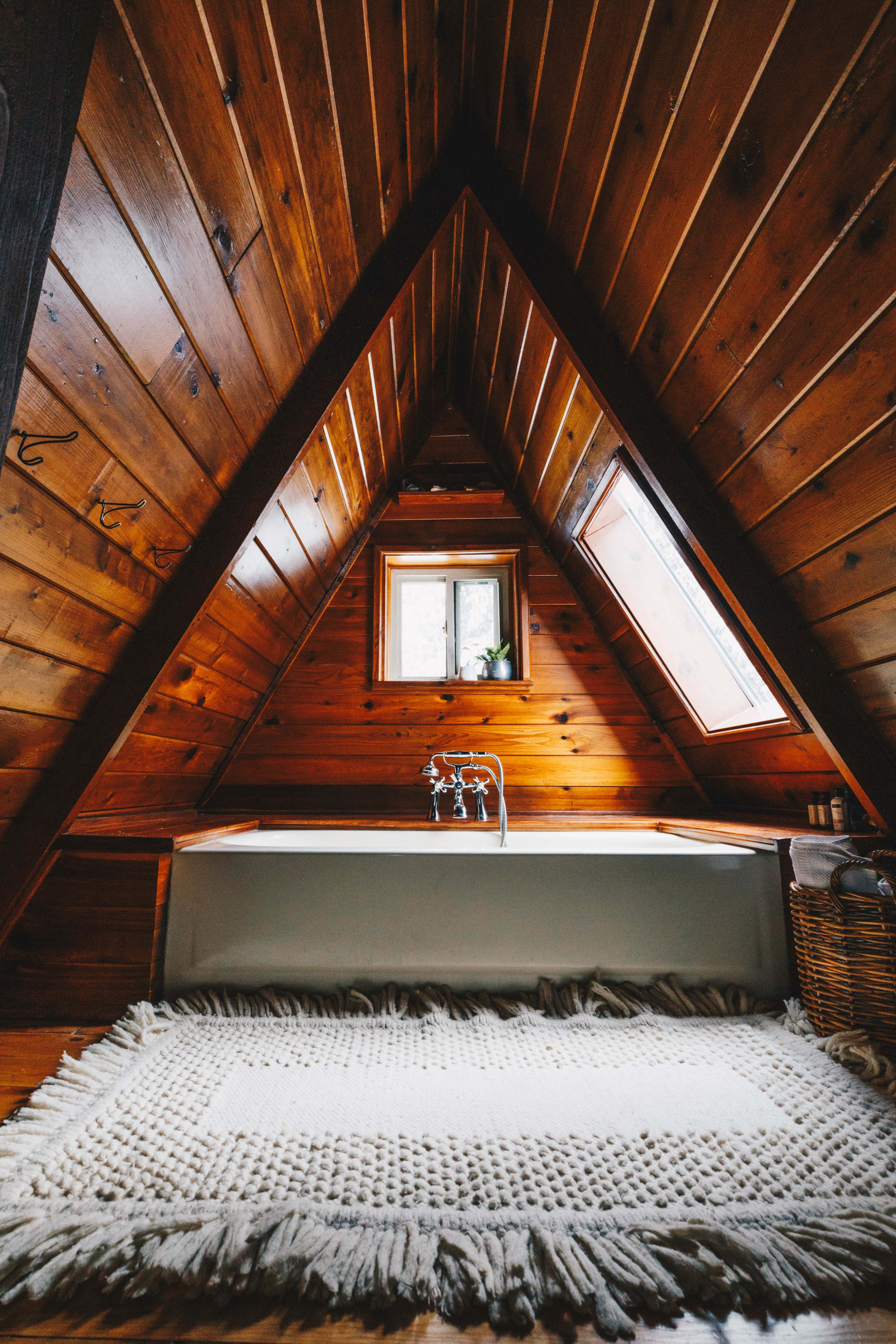 The image shows a triangular wooden bathroom with a freestanding bathtub and a window above it, set against a textured rug on the floor.