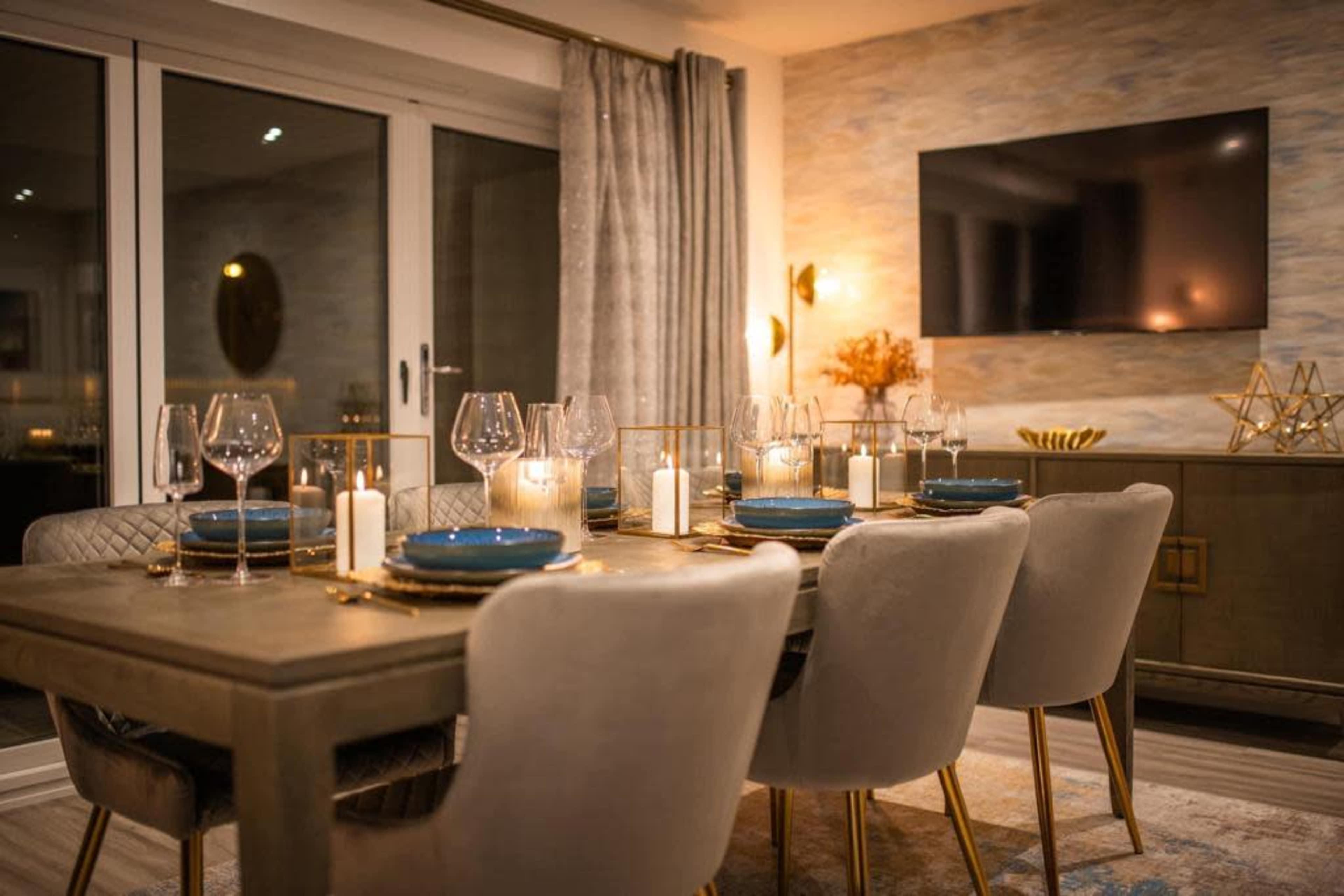 A modern dining room features a wooden table set with plates, glasses, and candles, accompanied by upholstered chairs and a TV mounted on the wall.