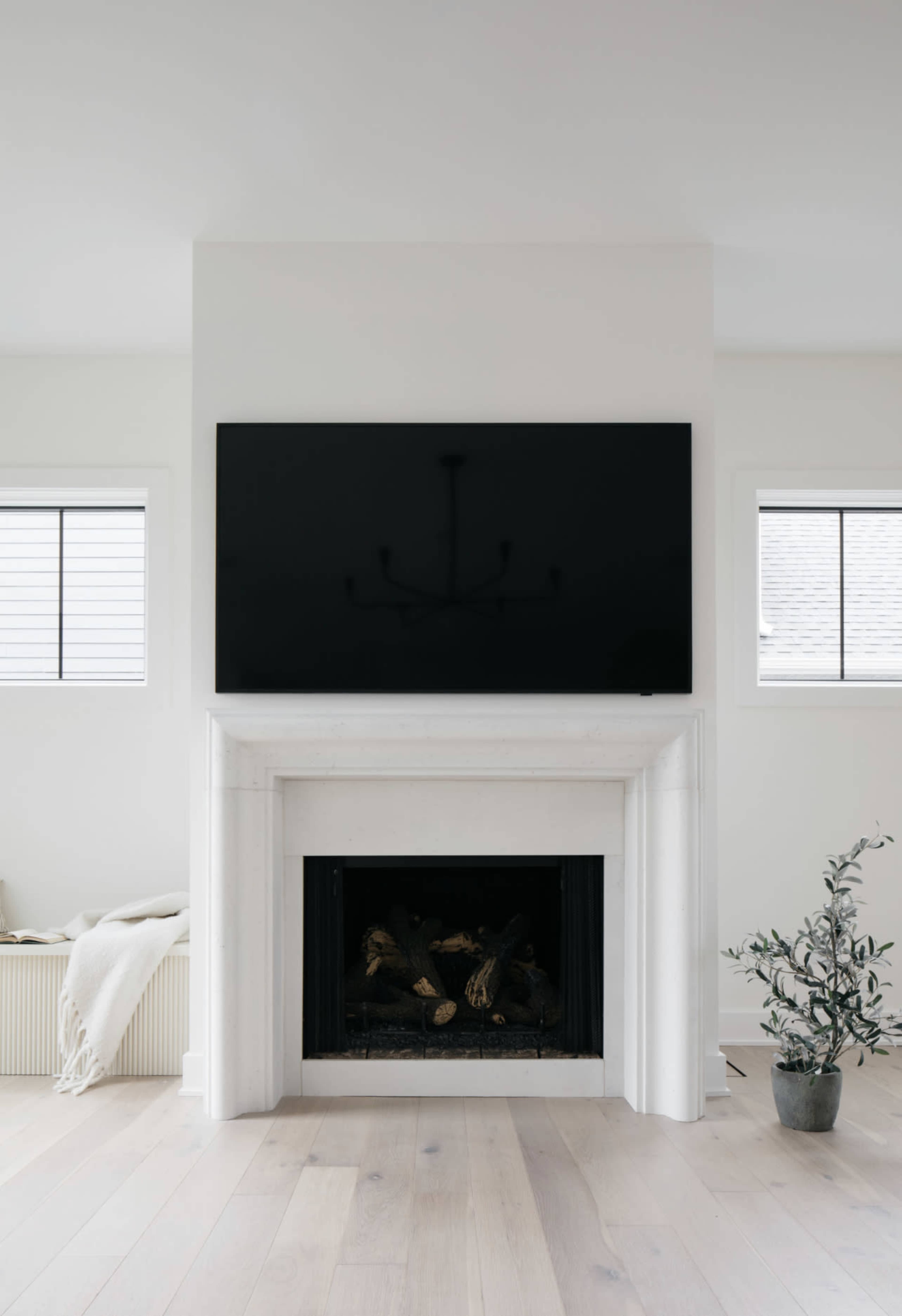 A modern living room features a white fireplace with a television mounted above it and large windows on either side.