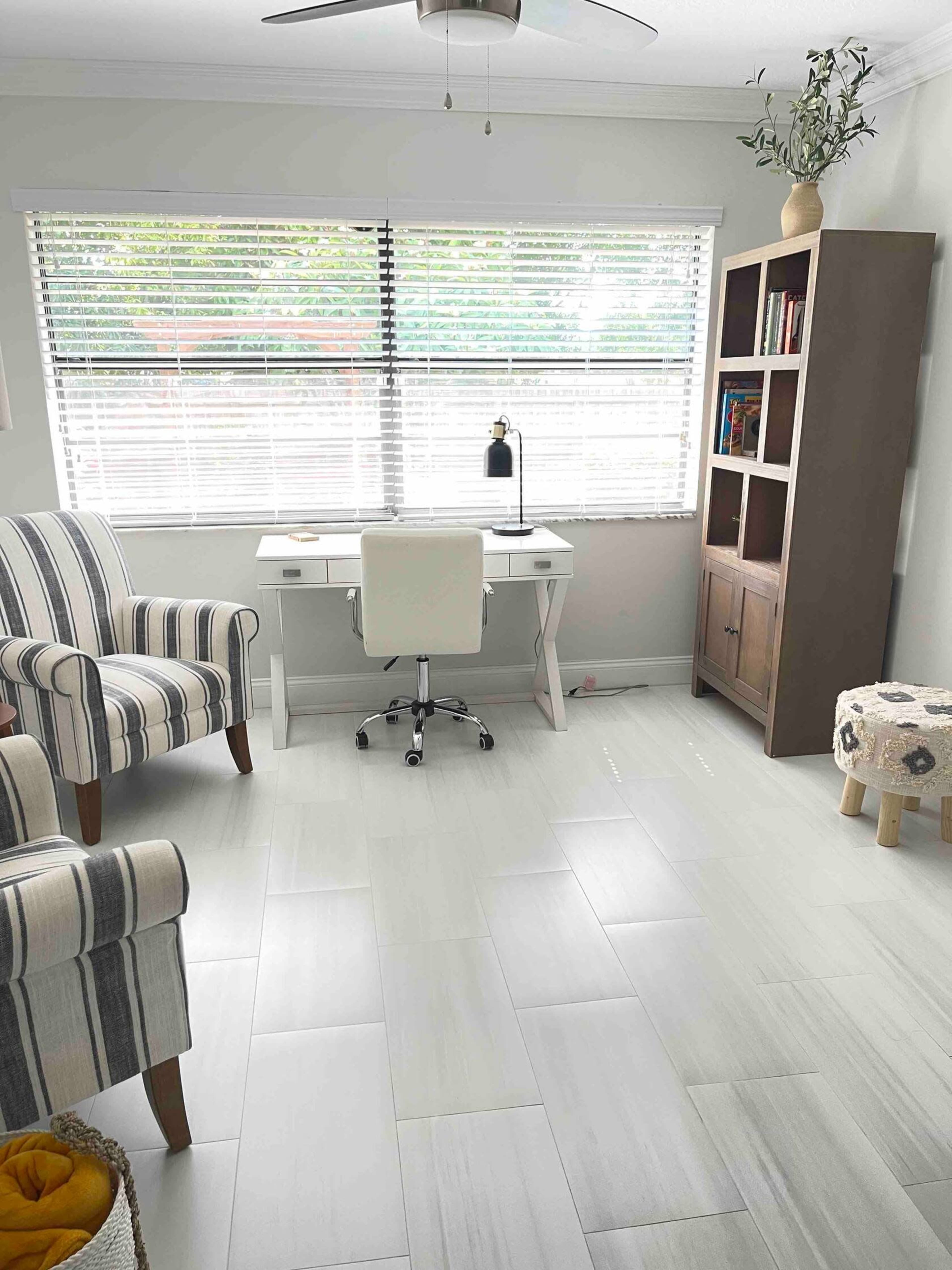 The image shows a well-lit office space with a desk, chair, bookshelf, and a striped armchair, arranged near a window with blinds.