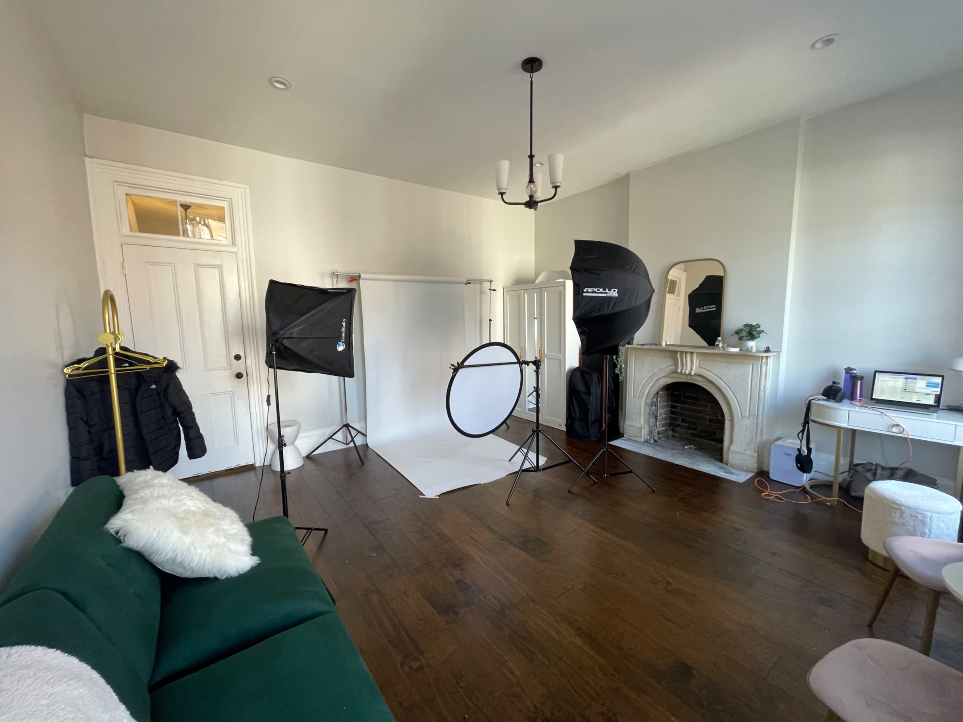 A well-lit room set up for photography, featuring a green couch, backdrop stands, light modifiers, and a mirror near an ornate fireplace.