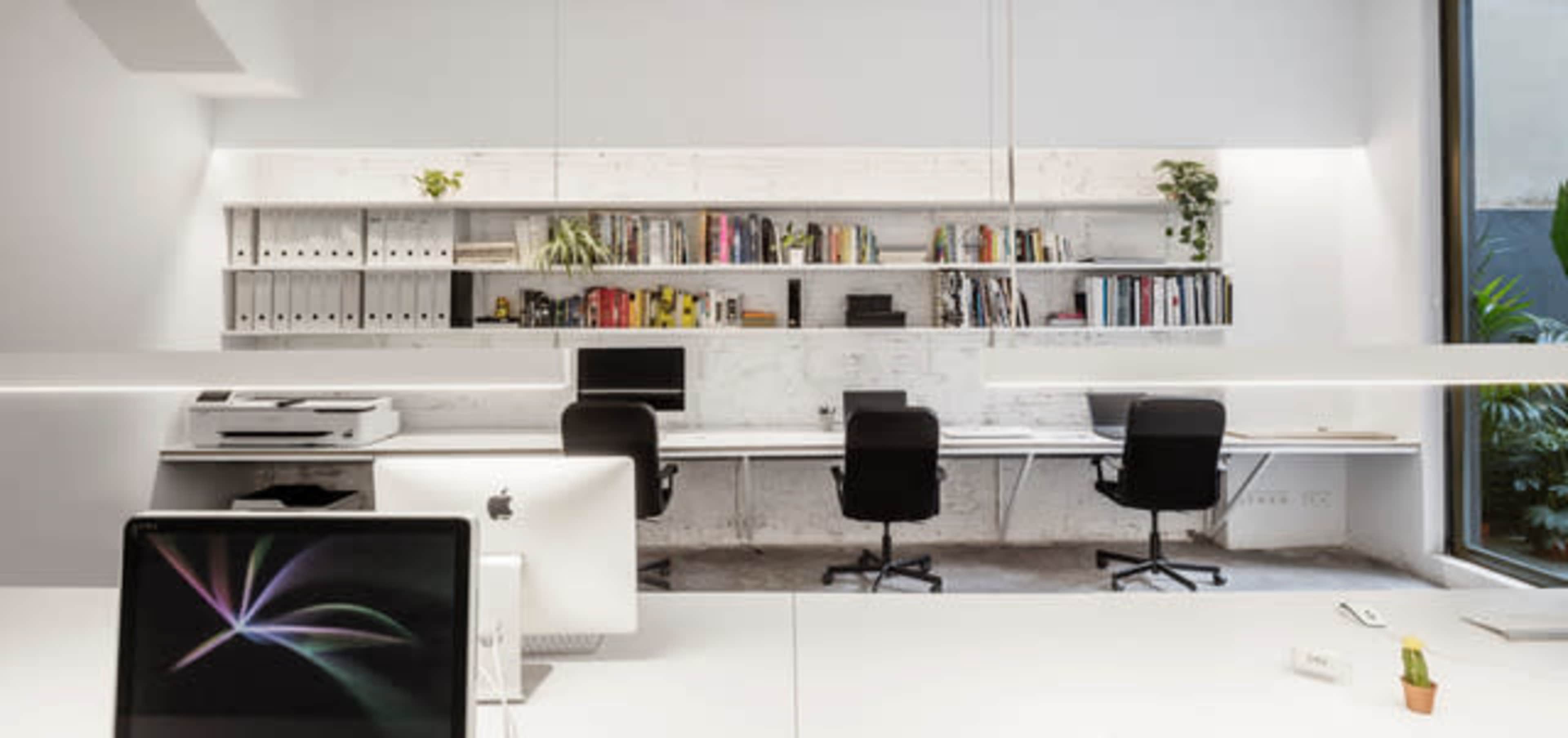 A modern office space with two black chairs at a long white desk, a row of bookshelves filled with books and binders, and a large window letting in natural light.