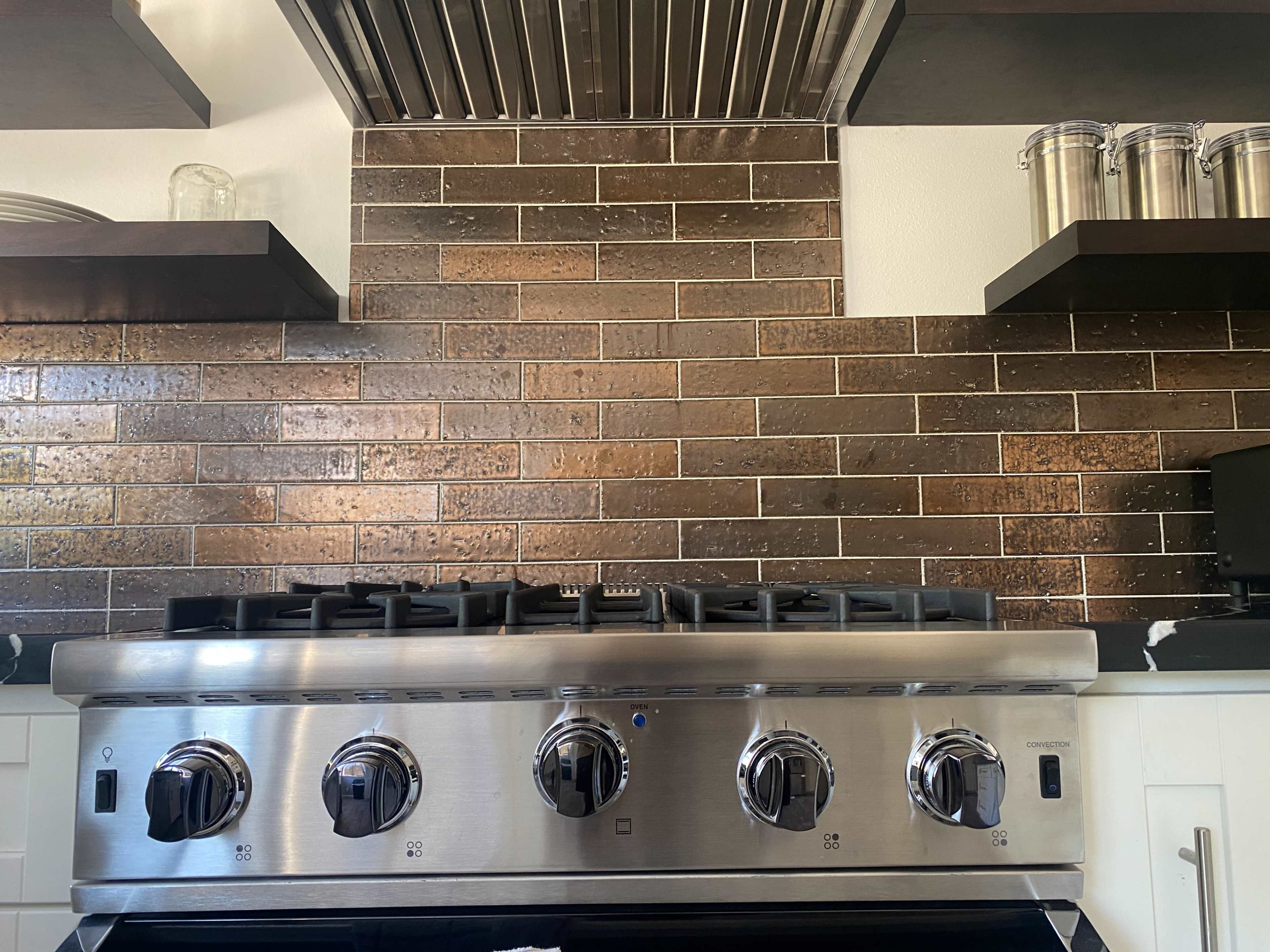 A stainless steel kitchen stove with four burners, set against a tiled backsplash and dark shelves holding glass containers.