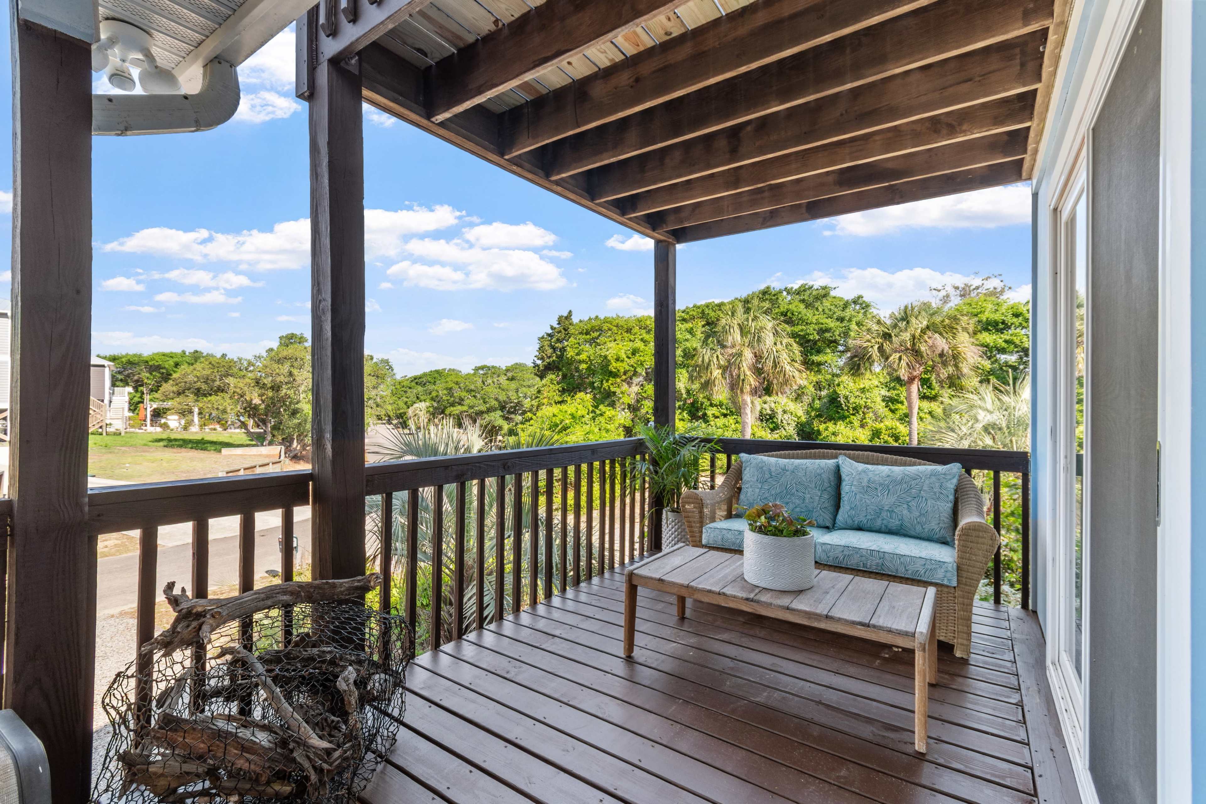 A wooden balcony features a small seating area with a potted plant and a view of lush greenery and blue skies.