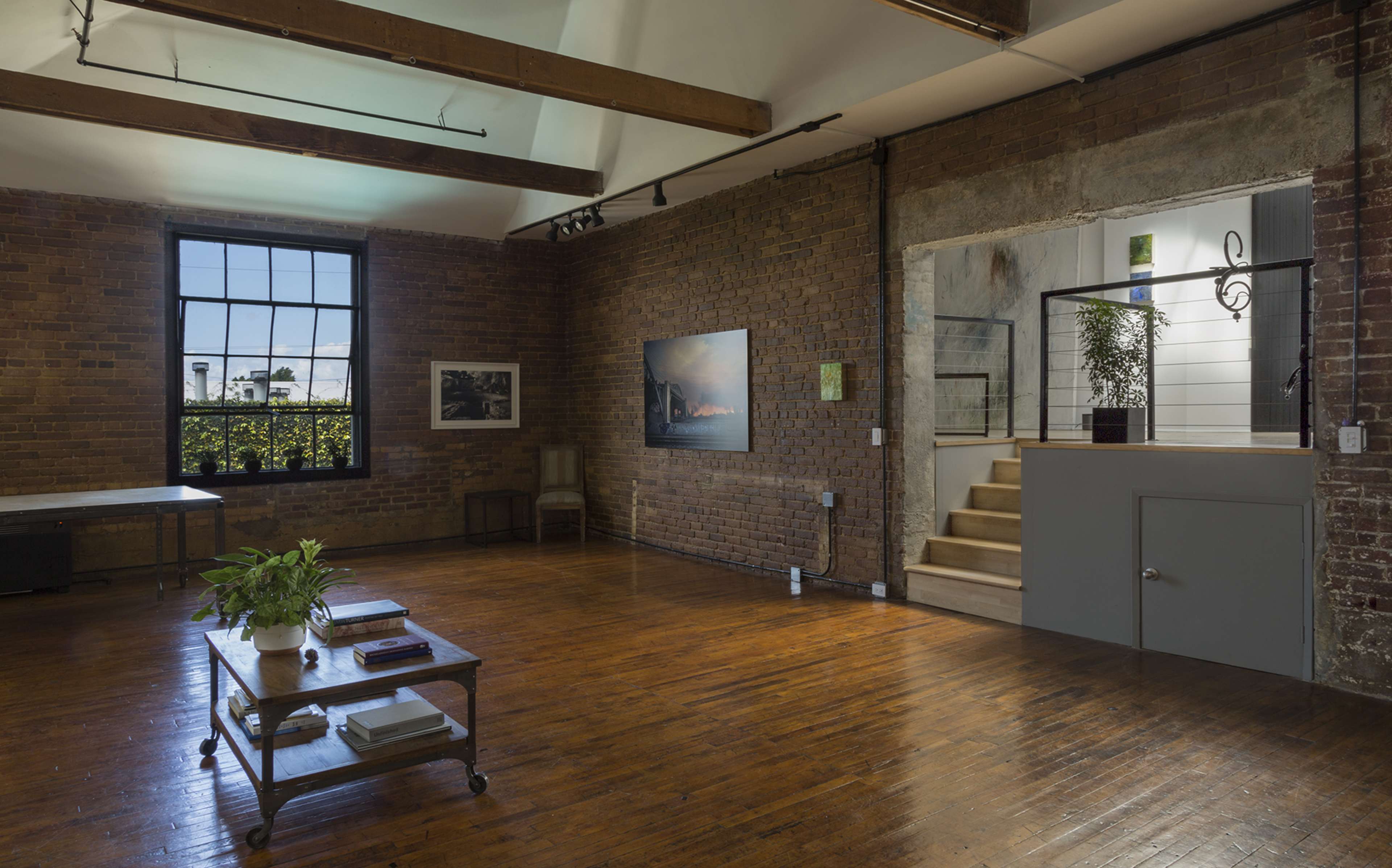 The interior of a spacious studio features exposed brick walls, wooden beams, and a set of stairs leading to a small alcove.