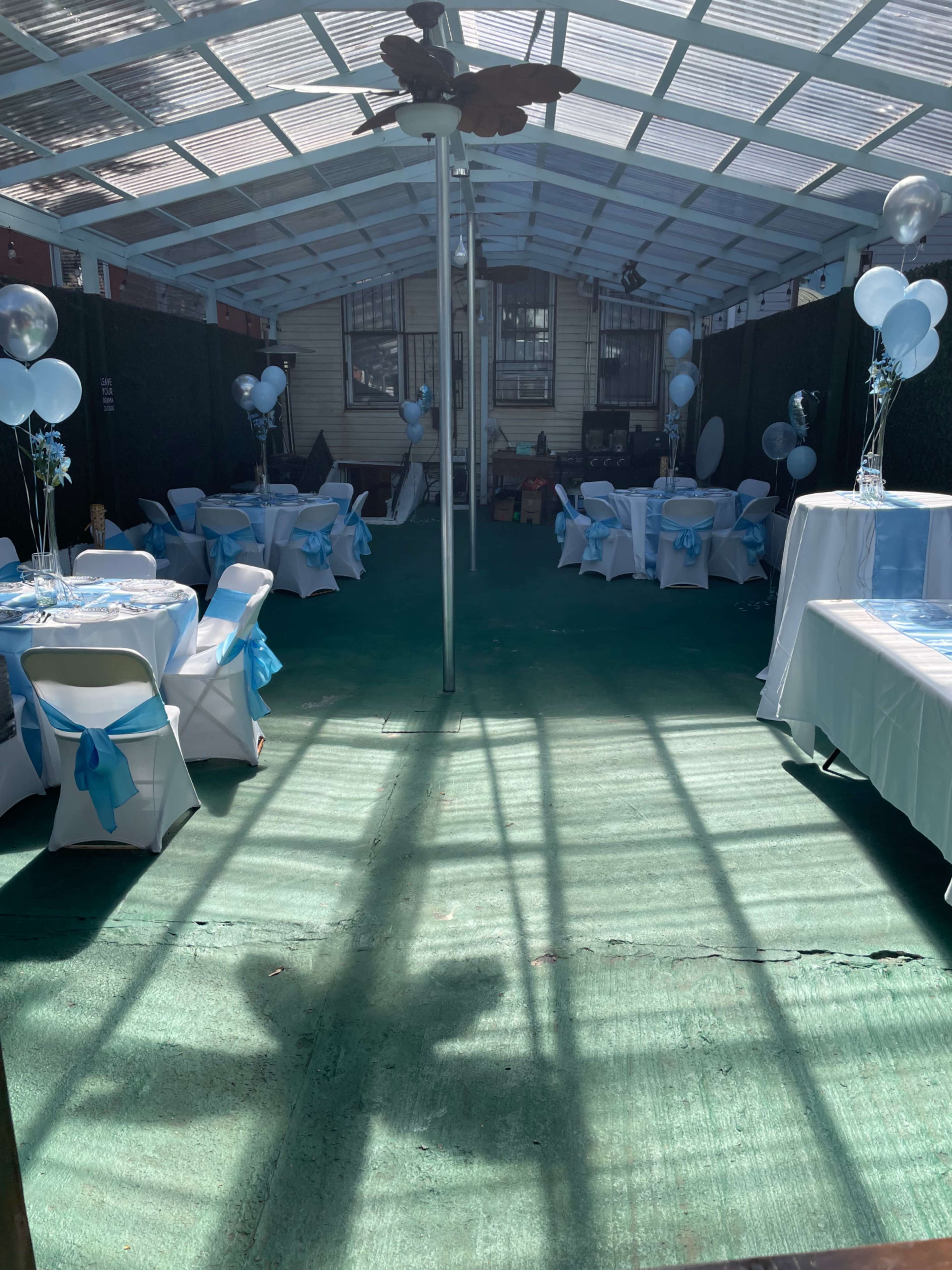 A decorated indoor space set up for an event, featuring tables covered in white cloths with blue accents and balloons.