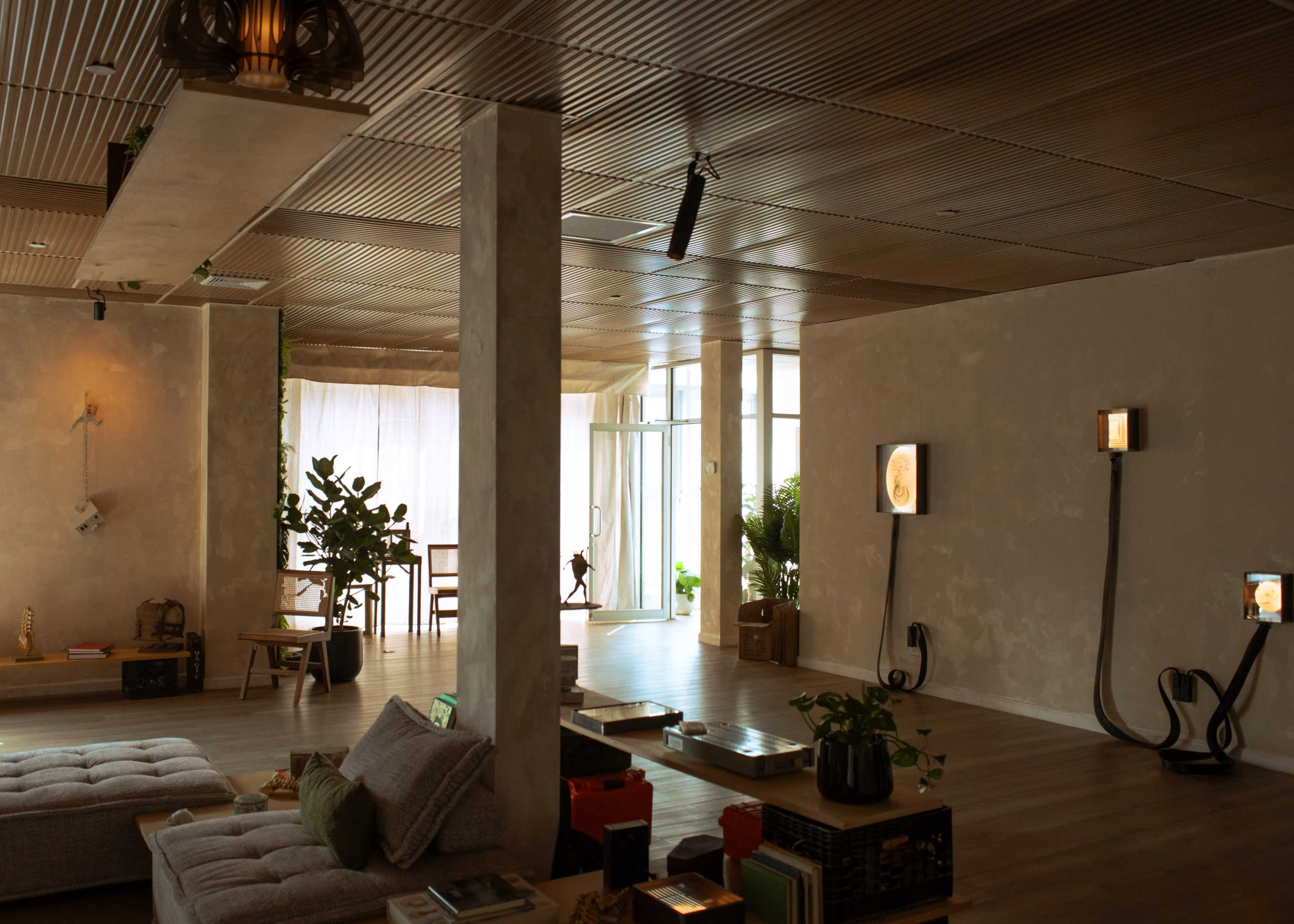 The image shows a spacious, modern living area with natural light, featuring minimalistic furniture, potted plants, and decorative wall-mounted elements.