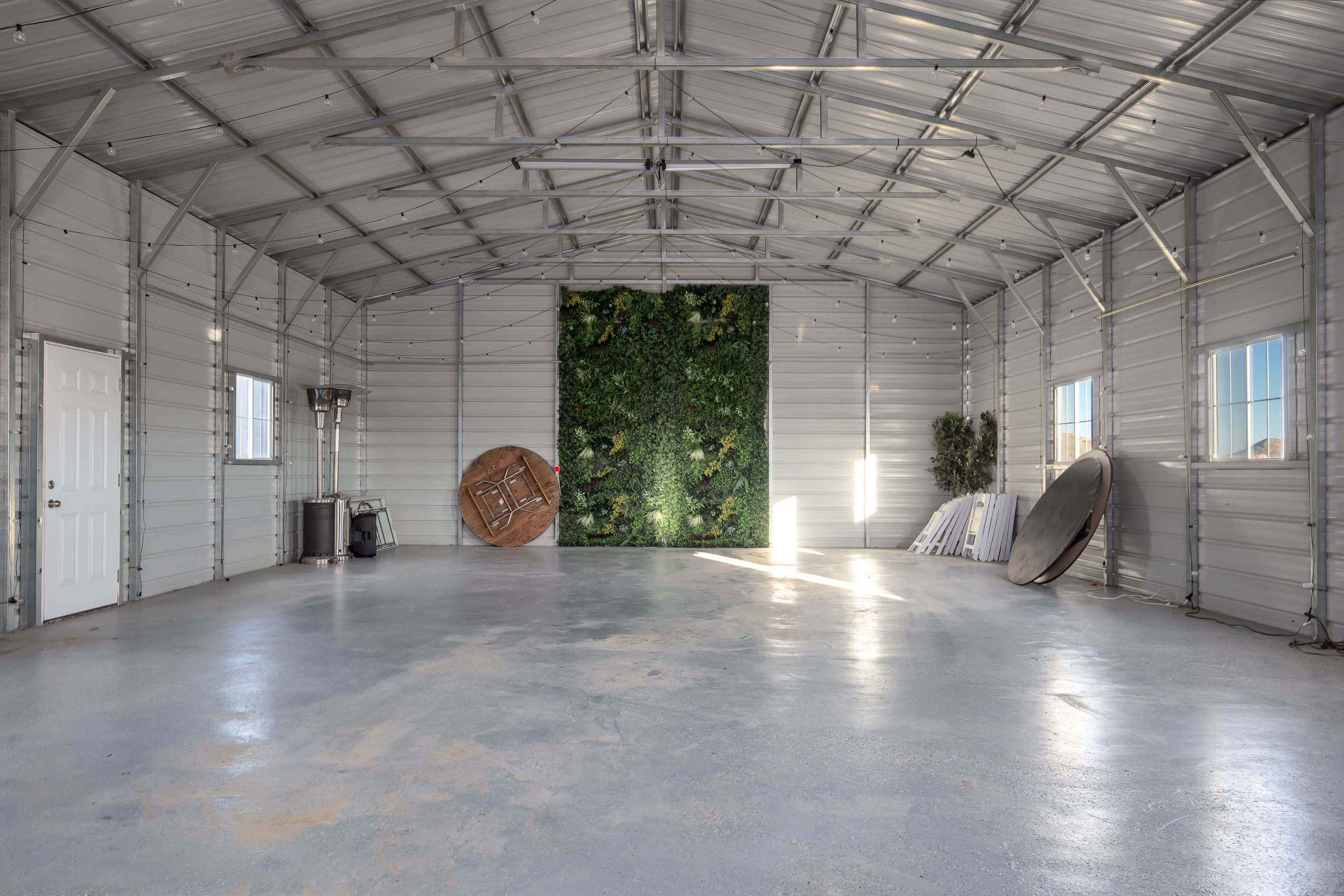 The interior of a large metal building features a green vertical plant wall, a large round wooden element, and various other simple furnishings.