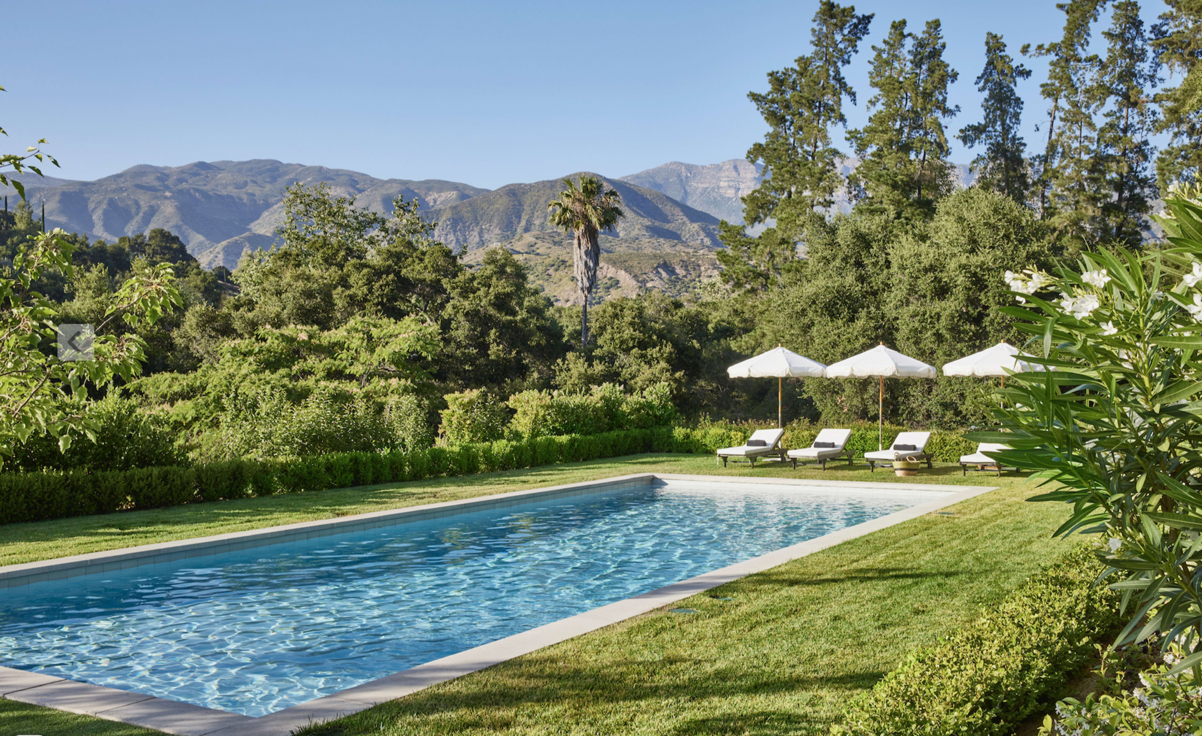 The image depicts a swimming pool surrounded by lush greenery and lounge chairs, with a backdrop of mountains under a clear blue sky.