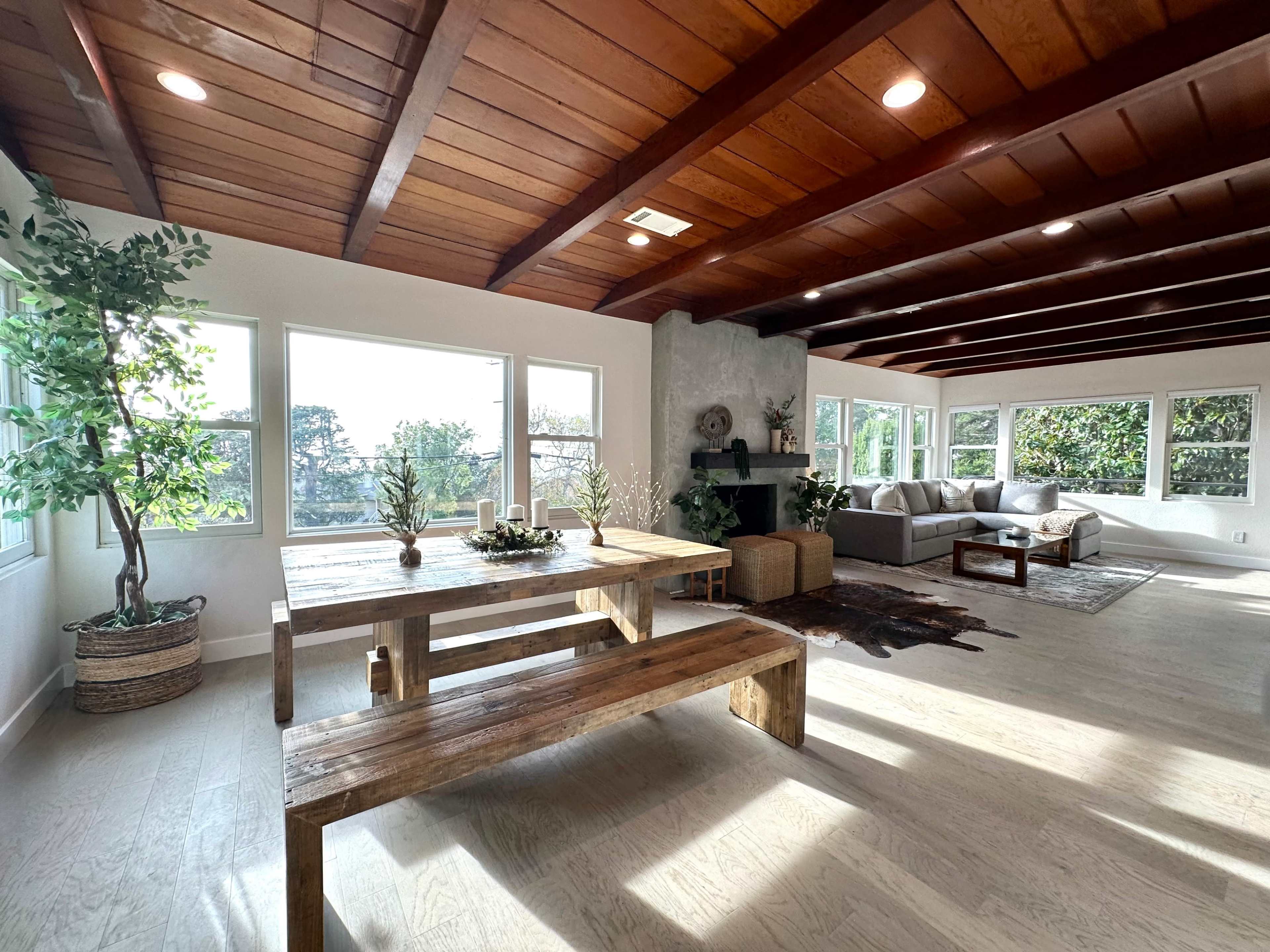 A bright, open living area featuring a wooden table with benches, a gray sofa, and large windows overlooking greenery.