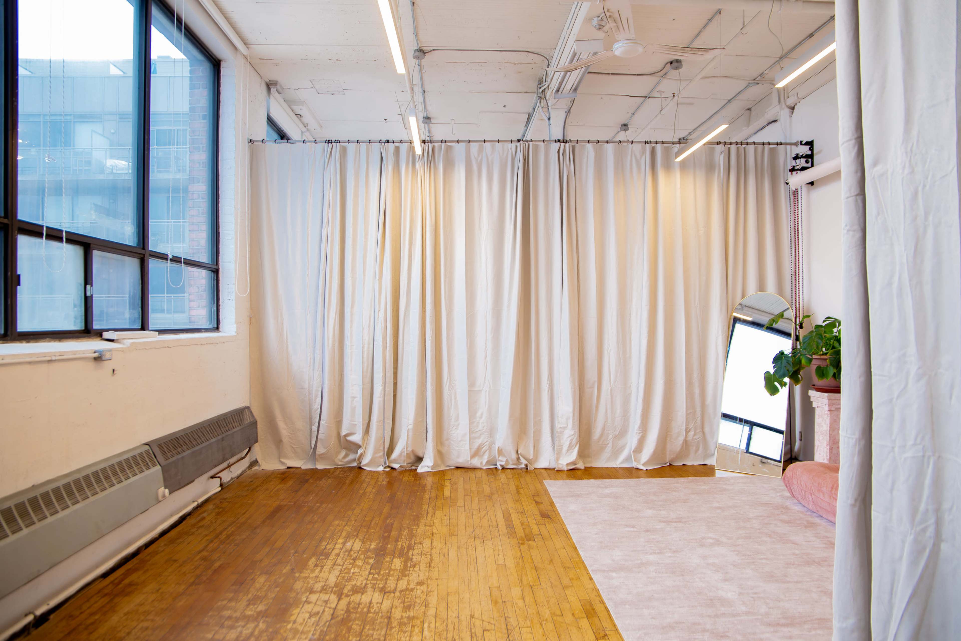 The image shows an empty room with large windows, light-colored curtains, a wooden floor, and a mirror leaning against a wall.