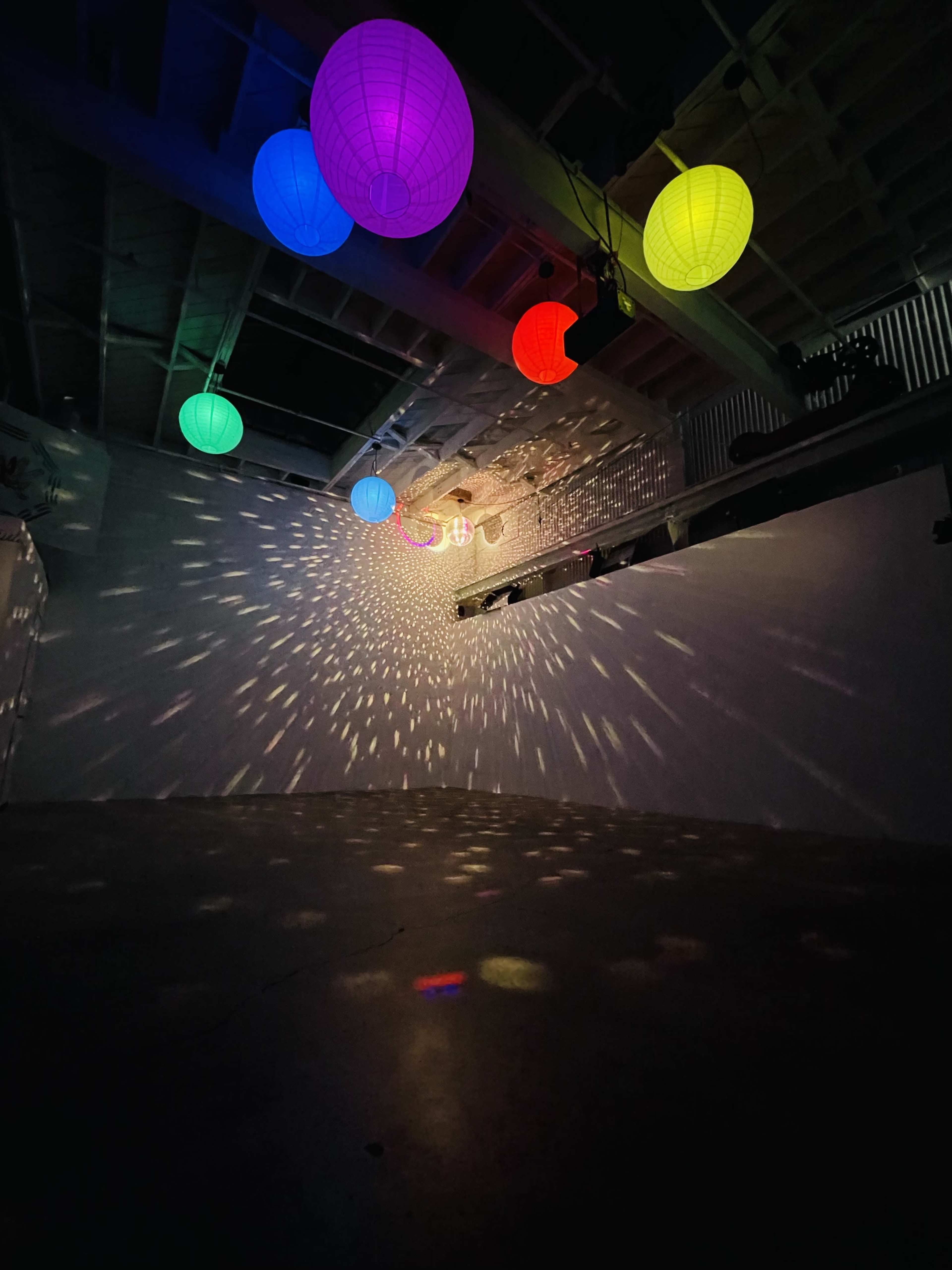 Colorful lanterns hang from the ceiling, casting patterned light across the floor and walls of a dimly lit room.