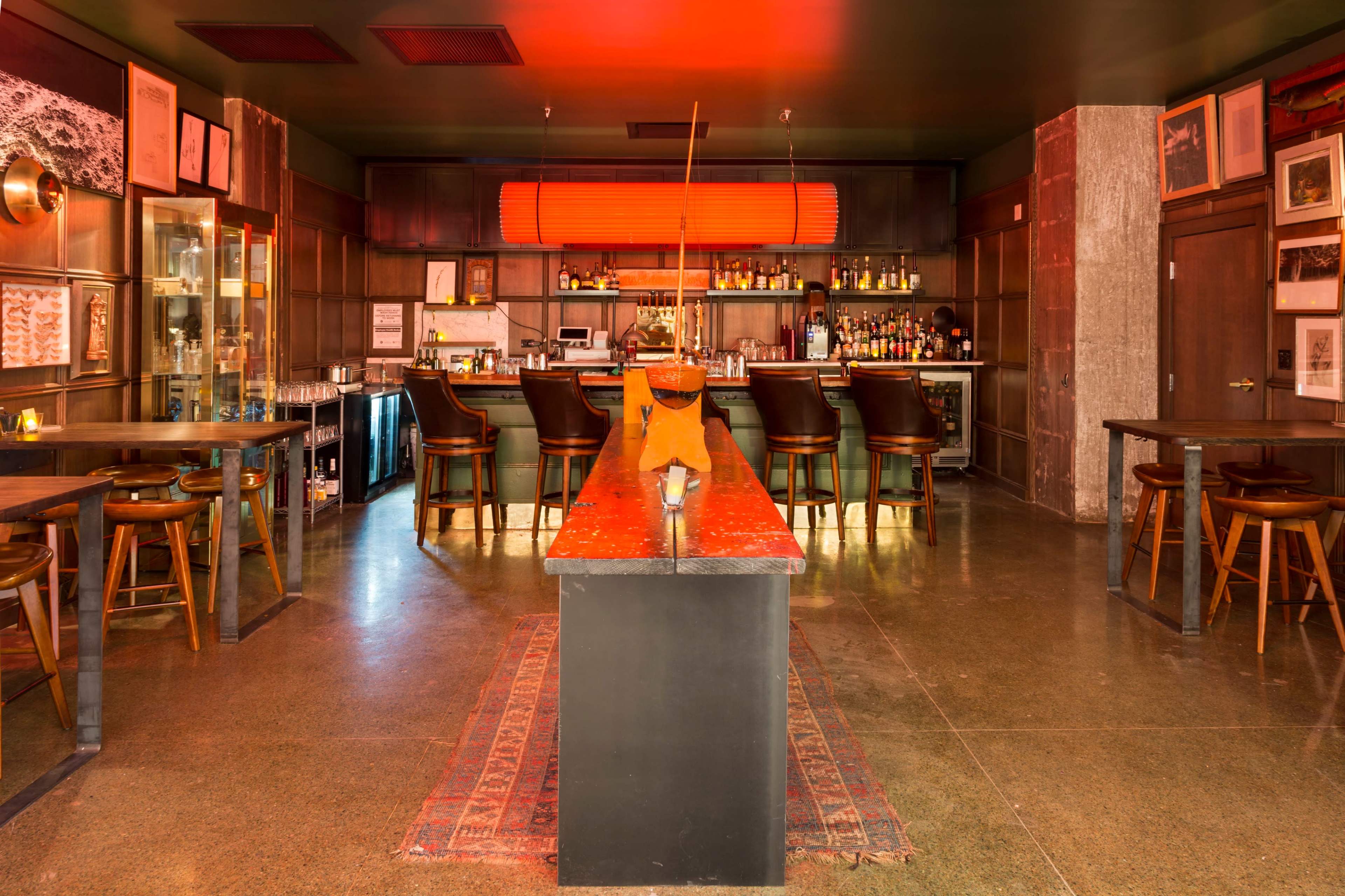 The image shows a modern bar with a long, dark countertop, wooden stools, and shelves stocked with various bottles, all illuminated by warm lighting.