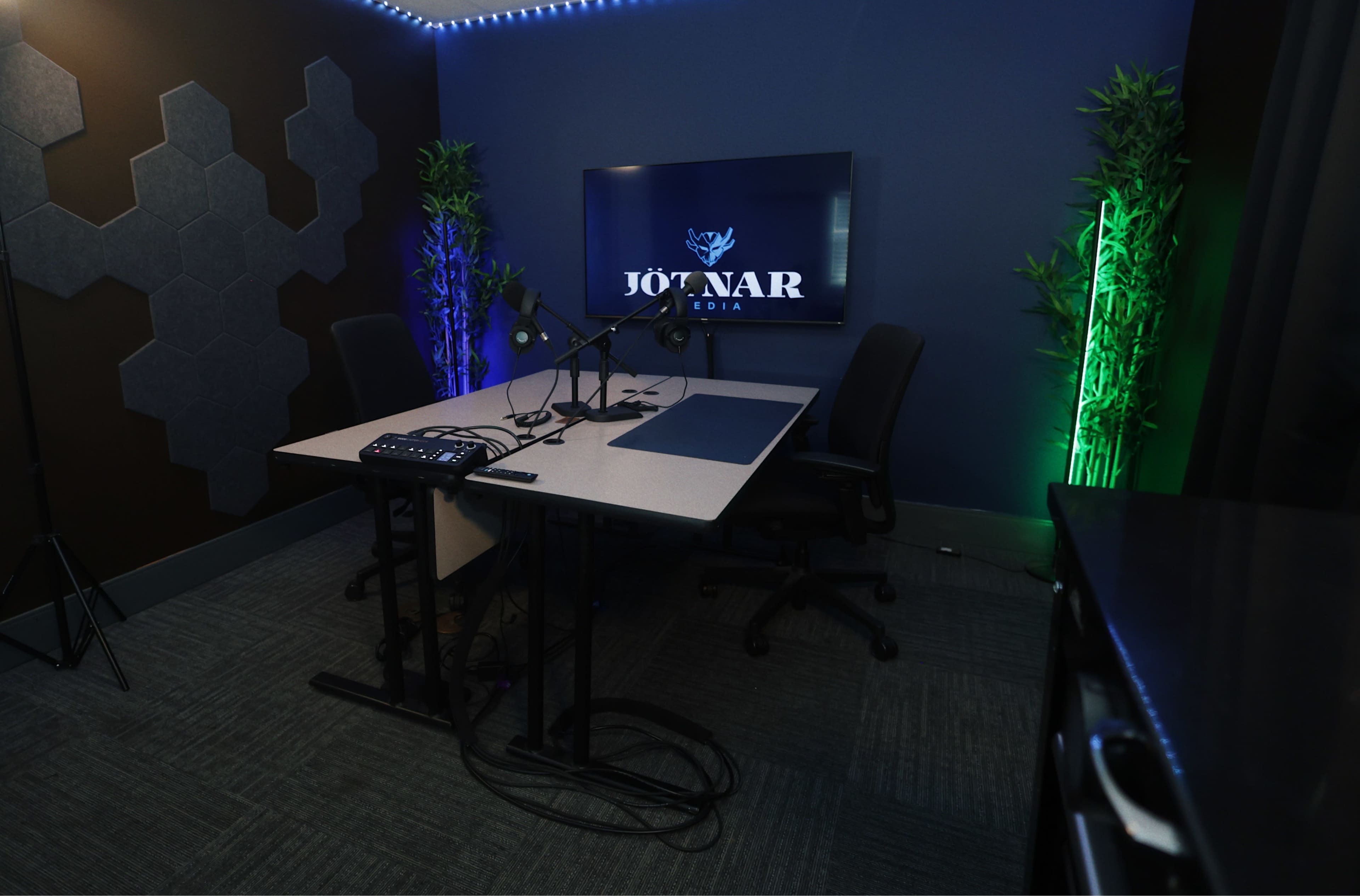 The image shows a modern podcast recording studio featuring a table with microphones, a large screen displaying the logo "JOTNAR MEDIA," and plants illuminated by colored lights.