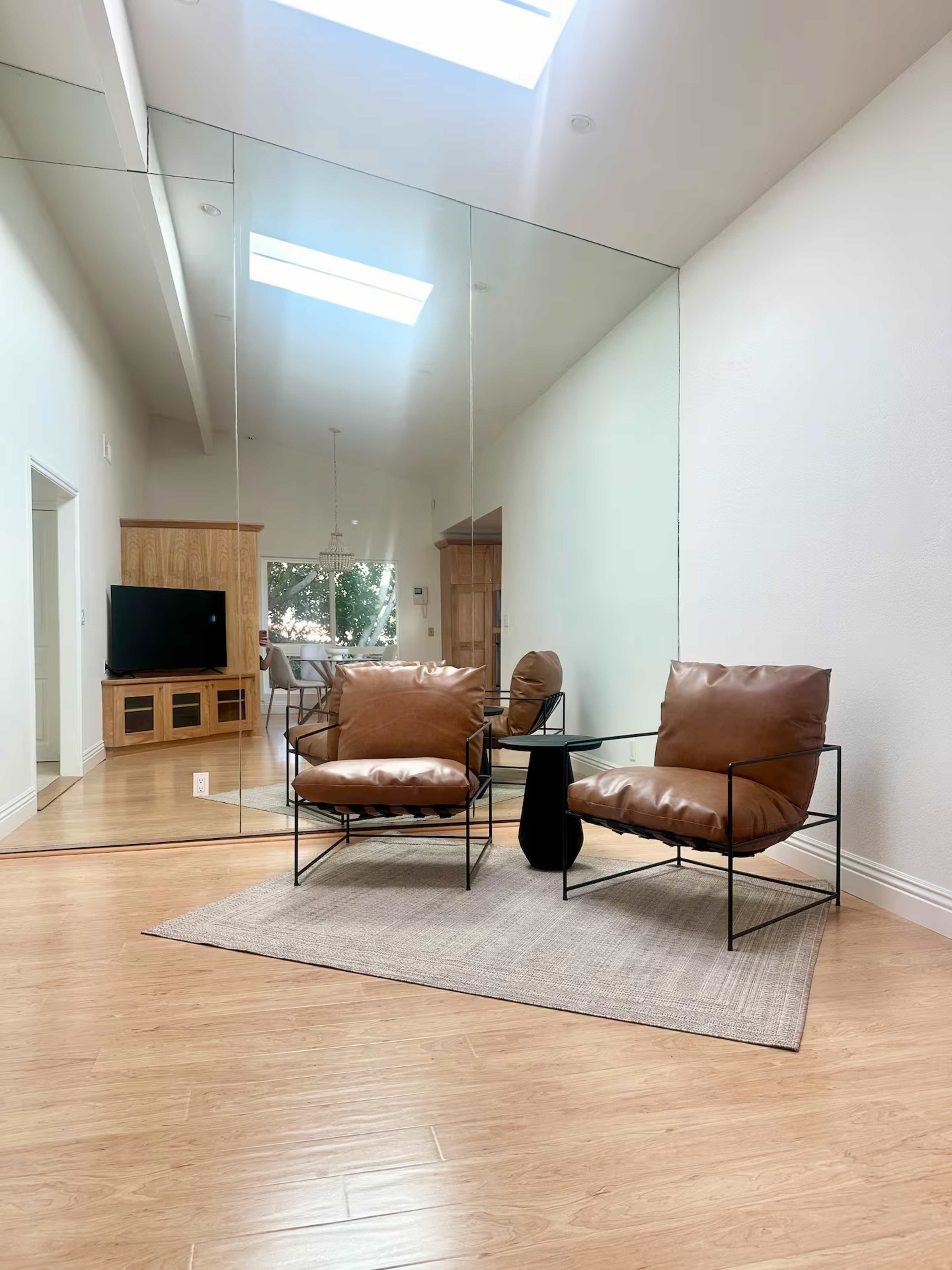 A spacious room features two brown leather chairs on a light rug, with a small black table between them, and a large mirror reflecting the space.
