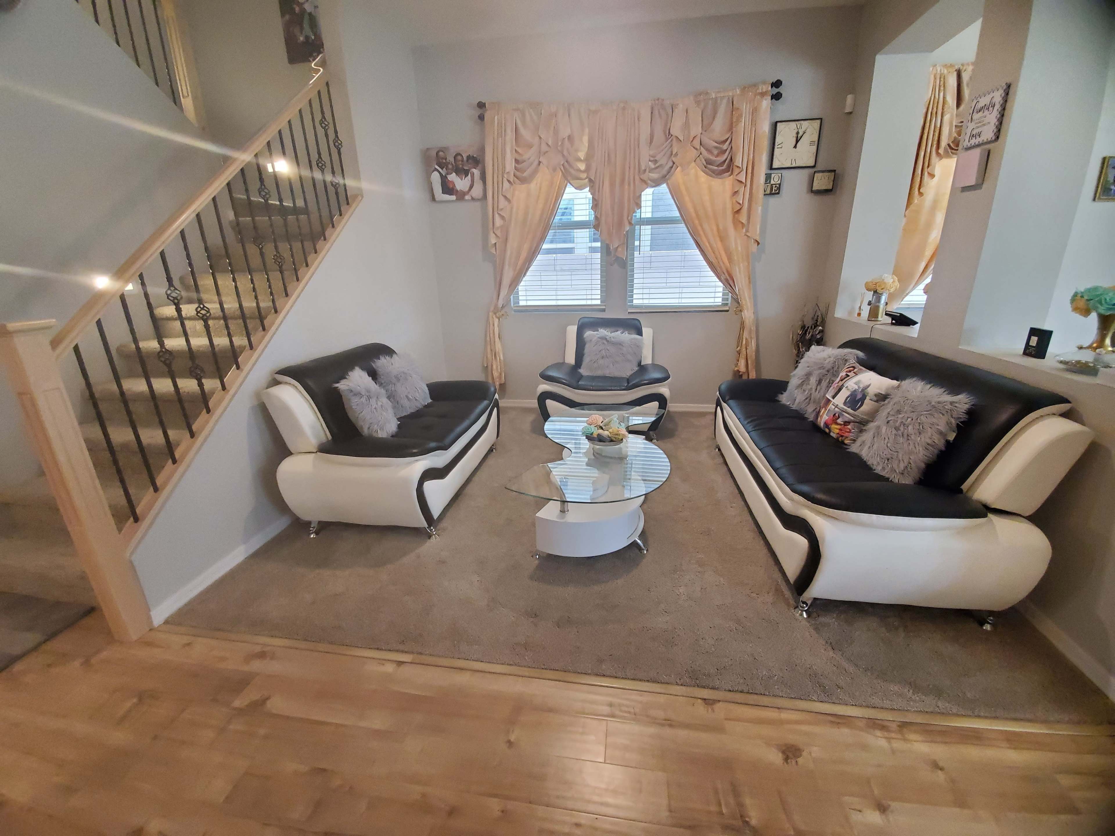 A living room with two modern black and white sofas, a glass coffee table, and large windows with drapes, adjacent to a staircase with a wooden railing.