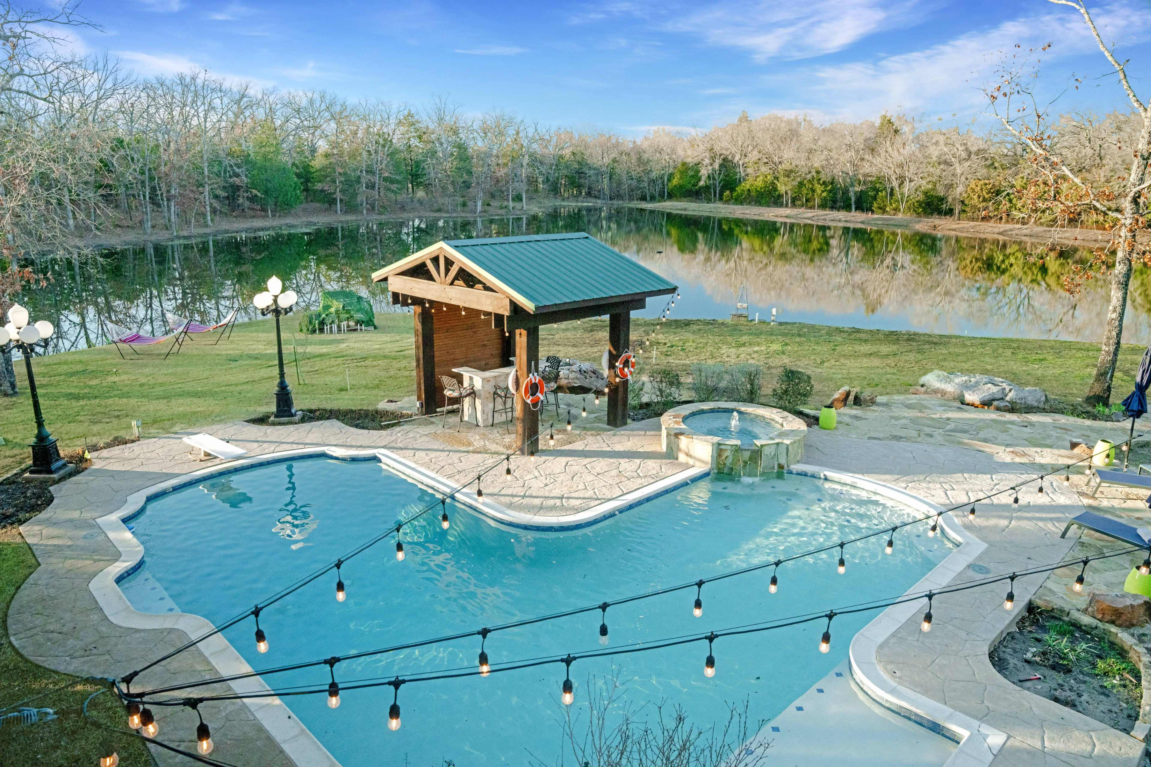 The image shows a pool area with a gazebo and a hot tub, surrounded by a landscaped yard and a lake in the background.