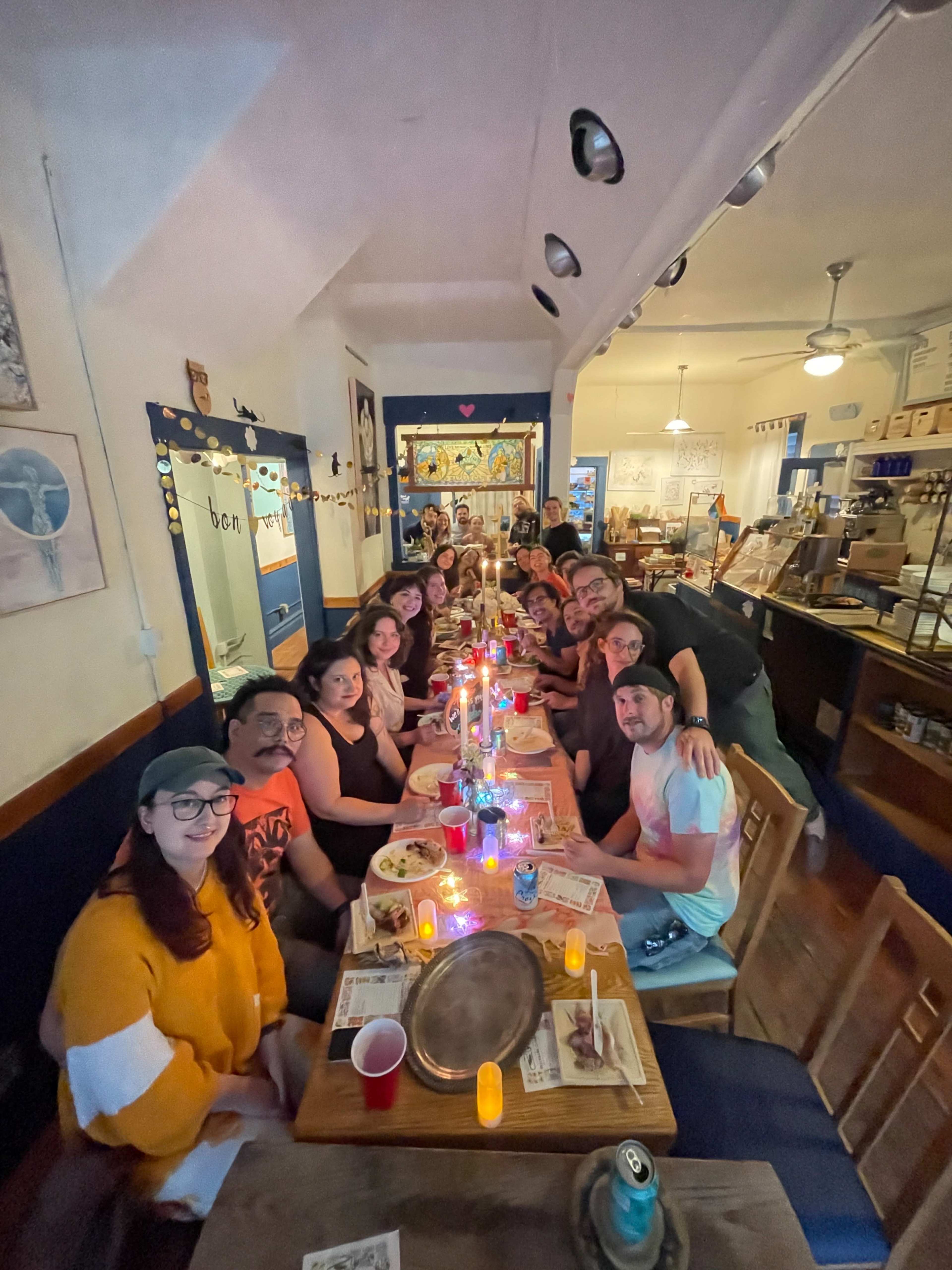 A group of people gathers around a long table in a brightly lit restaurant, sharing a meal with candles and drinks.
