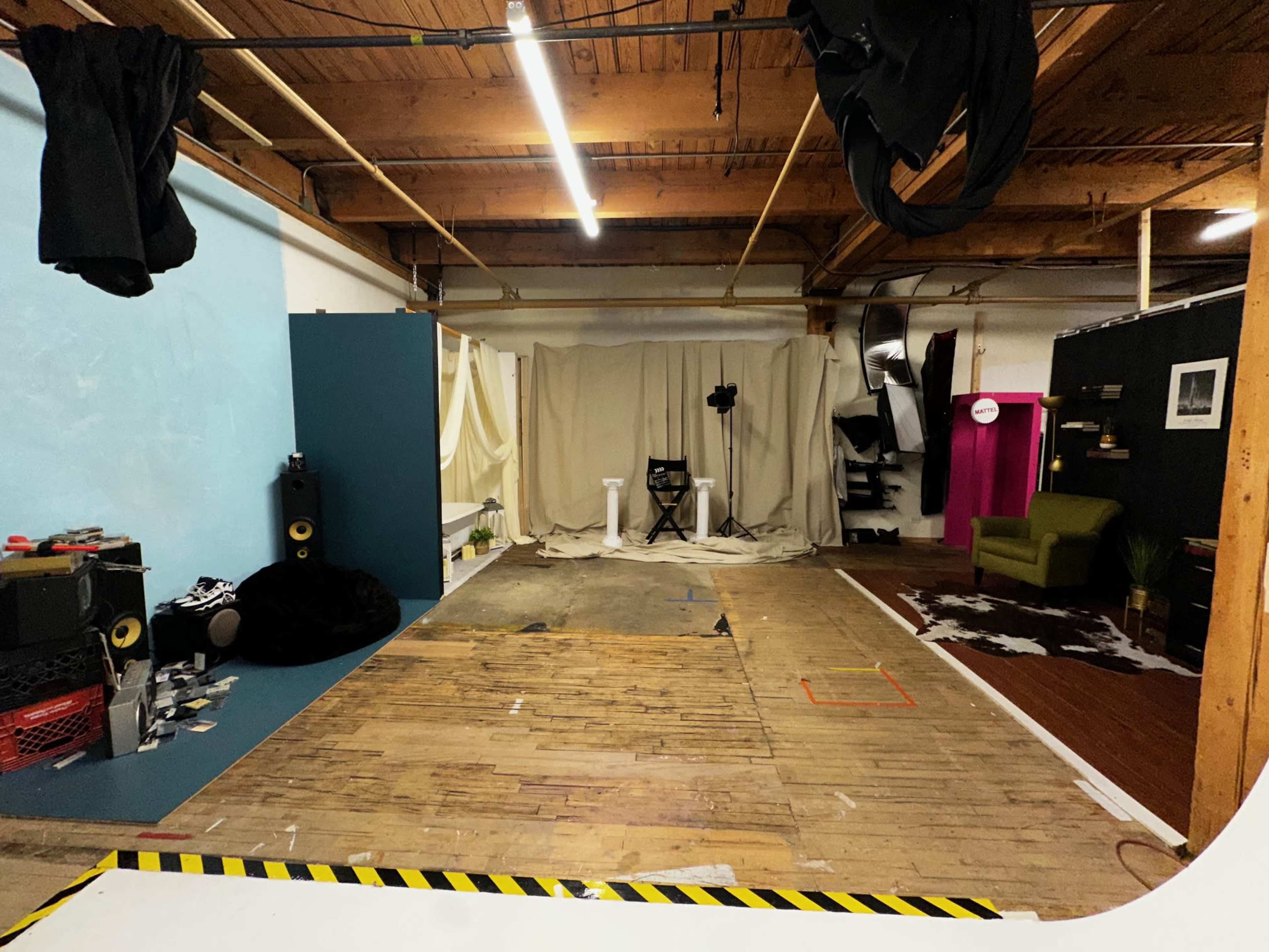 The image shows a spacious studio with wooden flooring, various backdrops, and scattered props for a photoshoot.