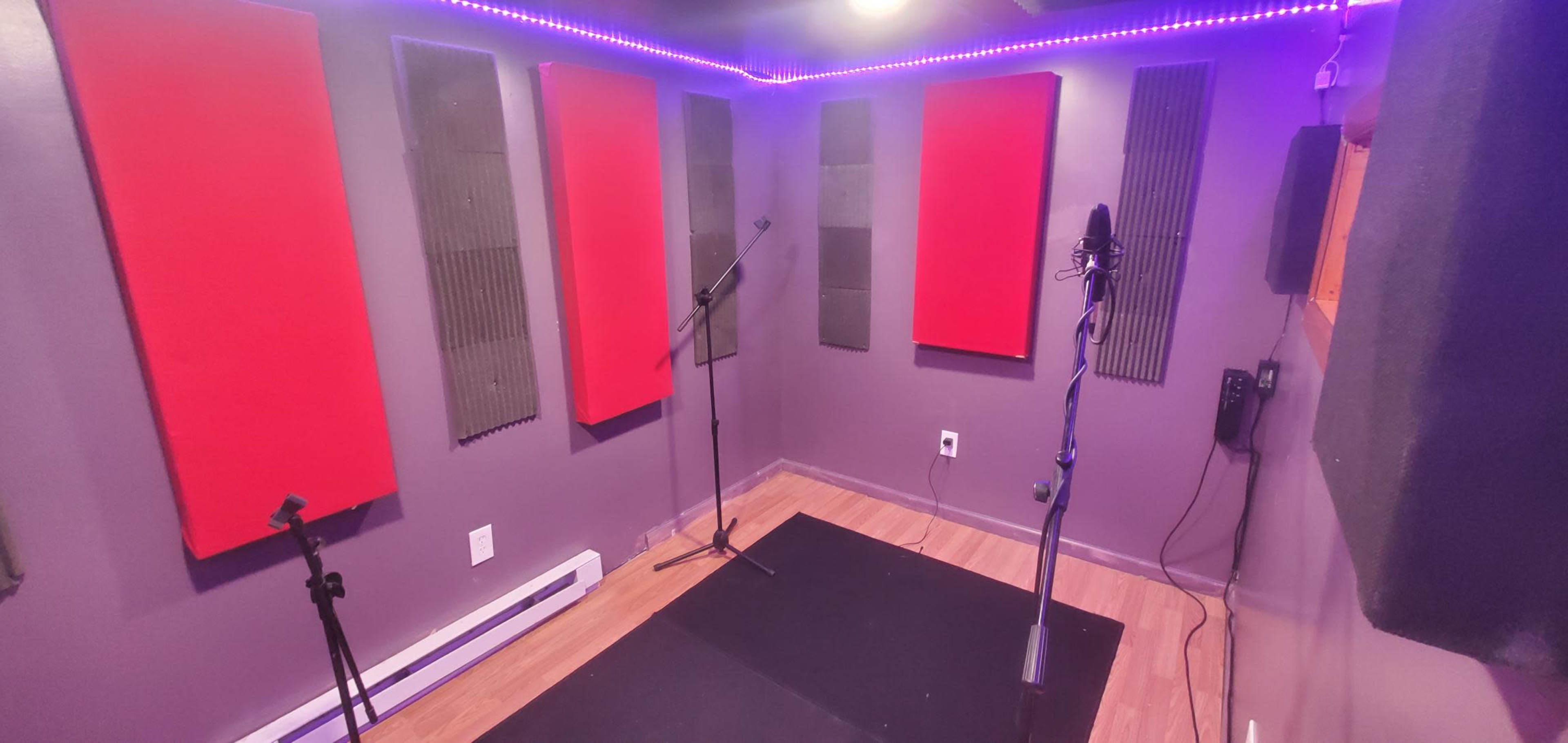 A small recording studio with red soundproofing panels, microphones on stands, and purple LED lighting.