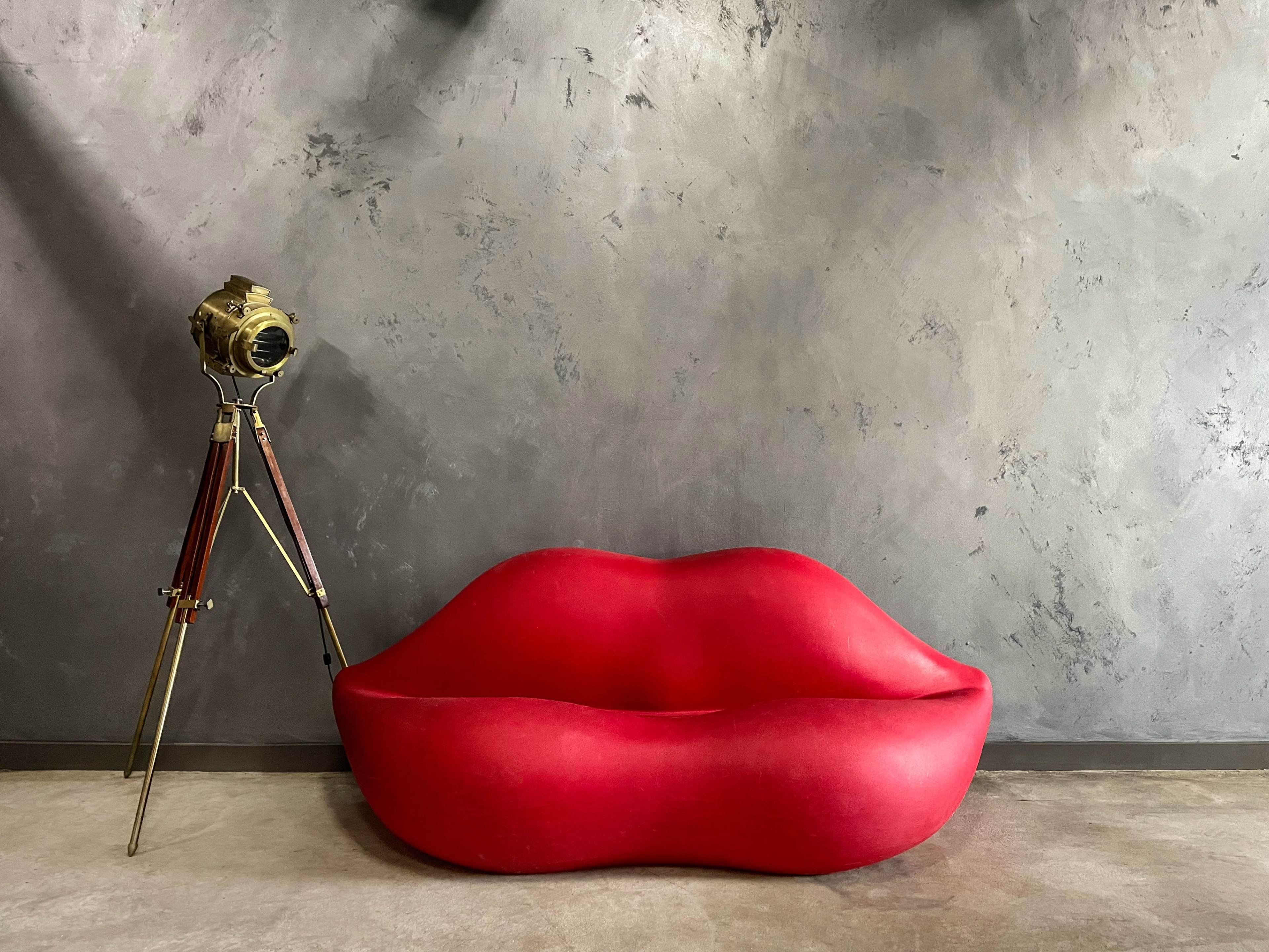 A red lip-shaped sofa is positioned next to a vintage-style camera on a tripod against a textured gray wall.