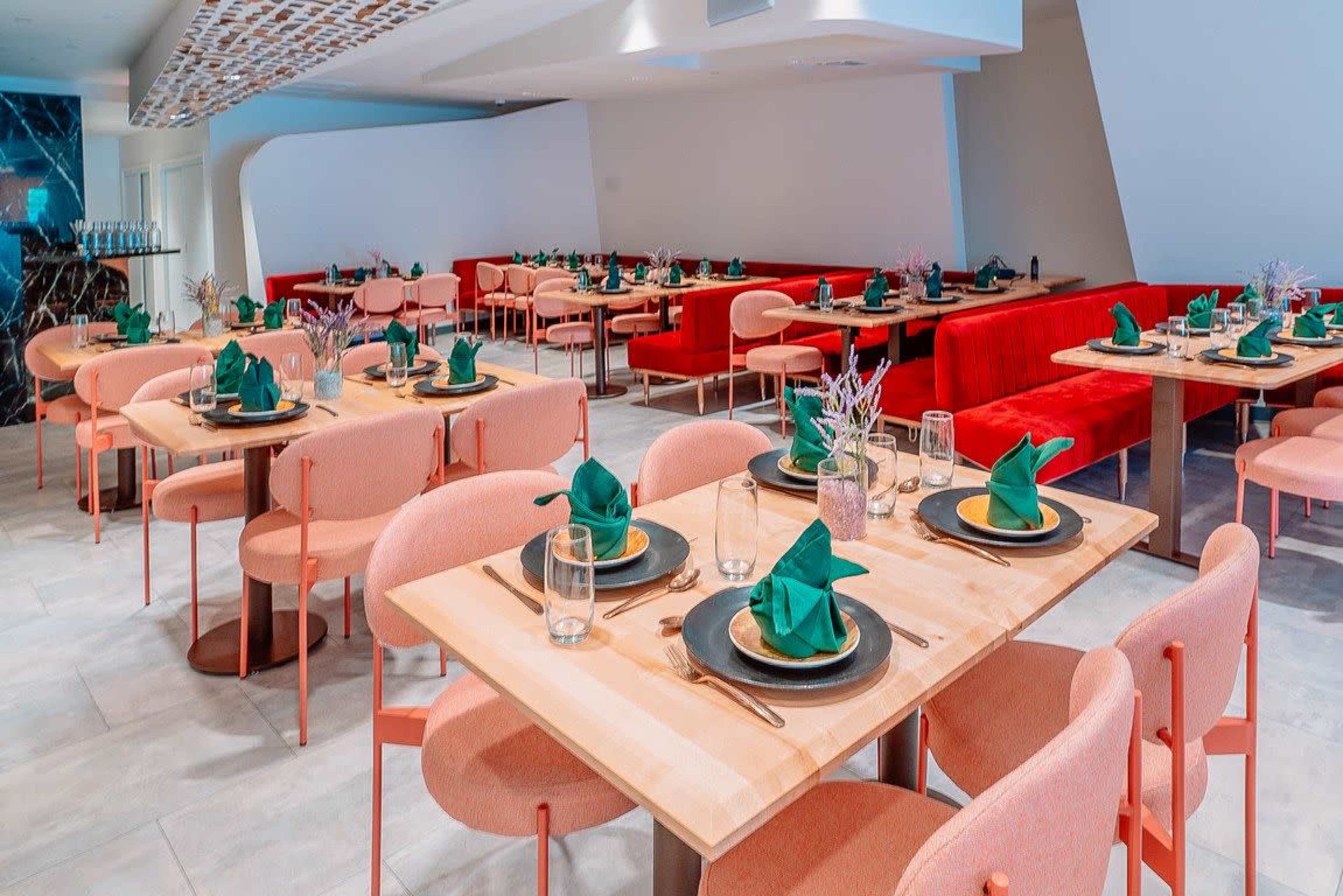 A brightly lit restaurant features several dining tables arranged with green napkins and black plates, surrounded by upholstered chairs in pink and red hues.