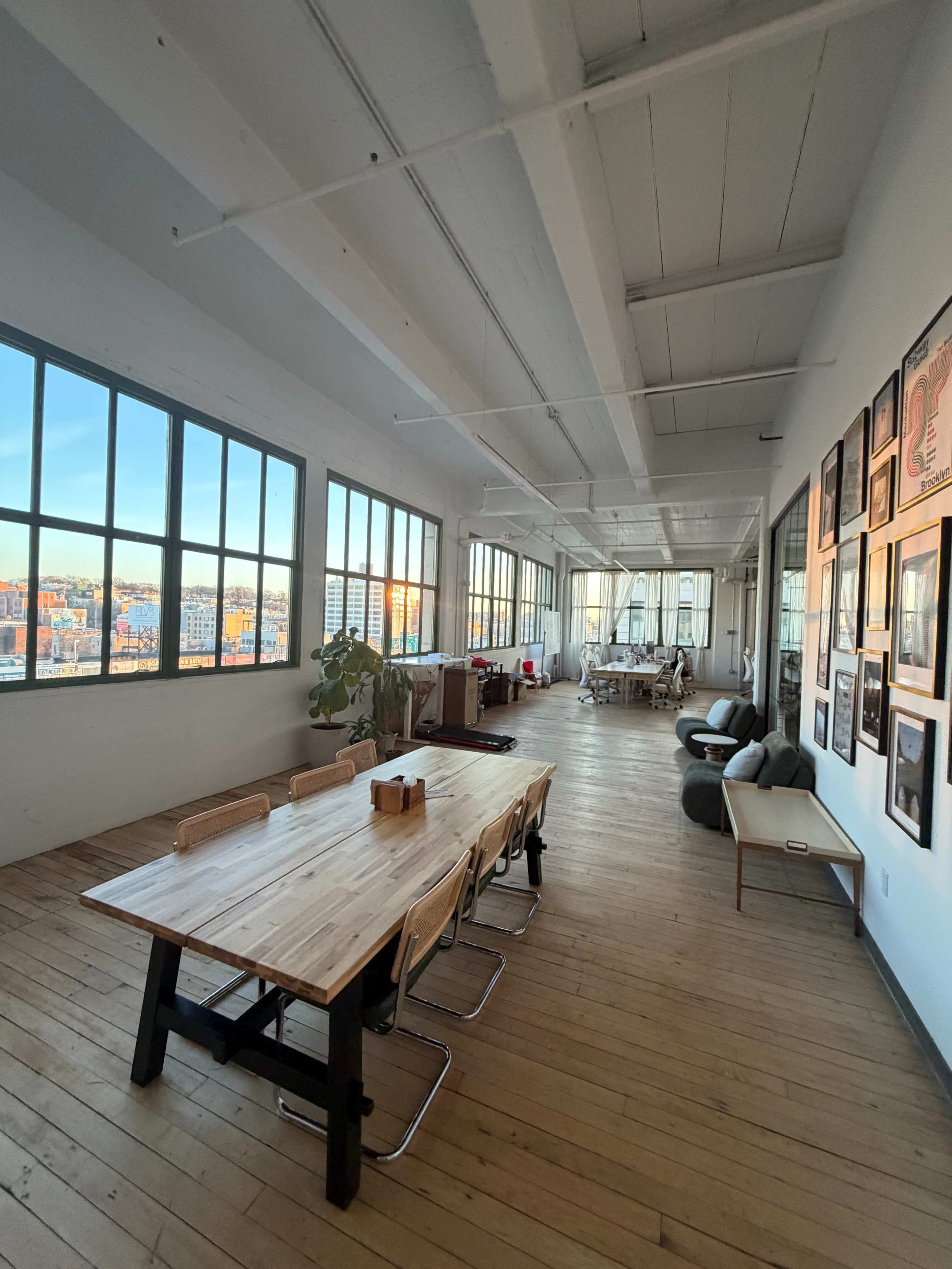 The image shows a spacious, well-lit office interior with large windows, wooden flooring, and a long wooden table surrounded by chairs.