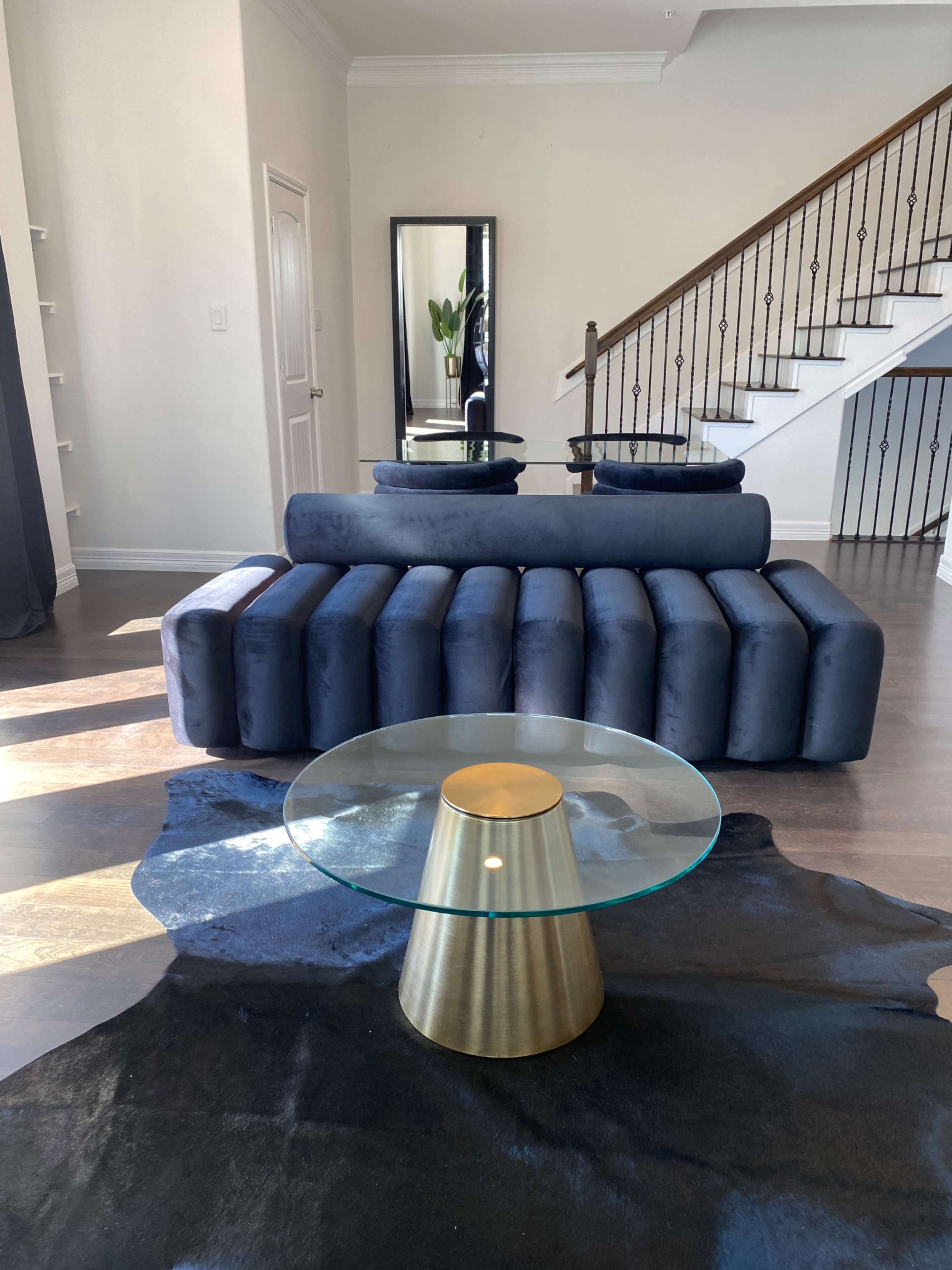 A blue, modern sofa with cylindrical sections sits in a living room alongside a glass-top coffee table and a decorative rug, against a backdrop of a staircase and a mirror.