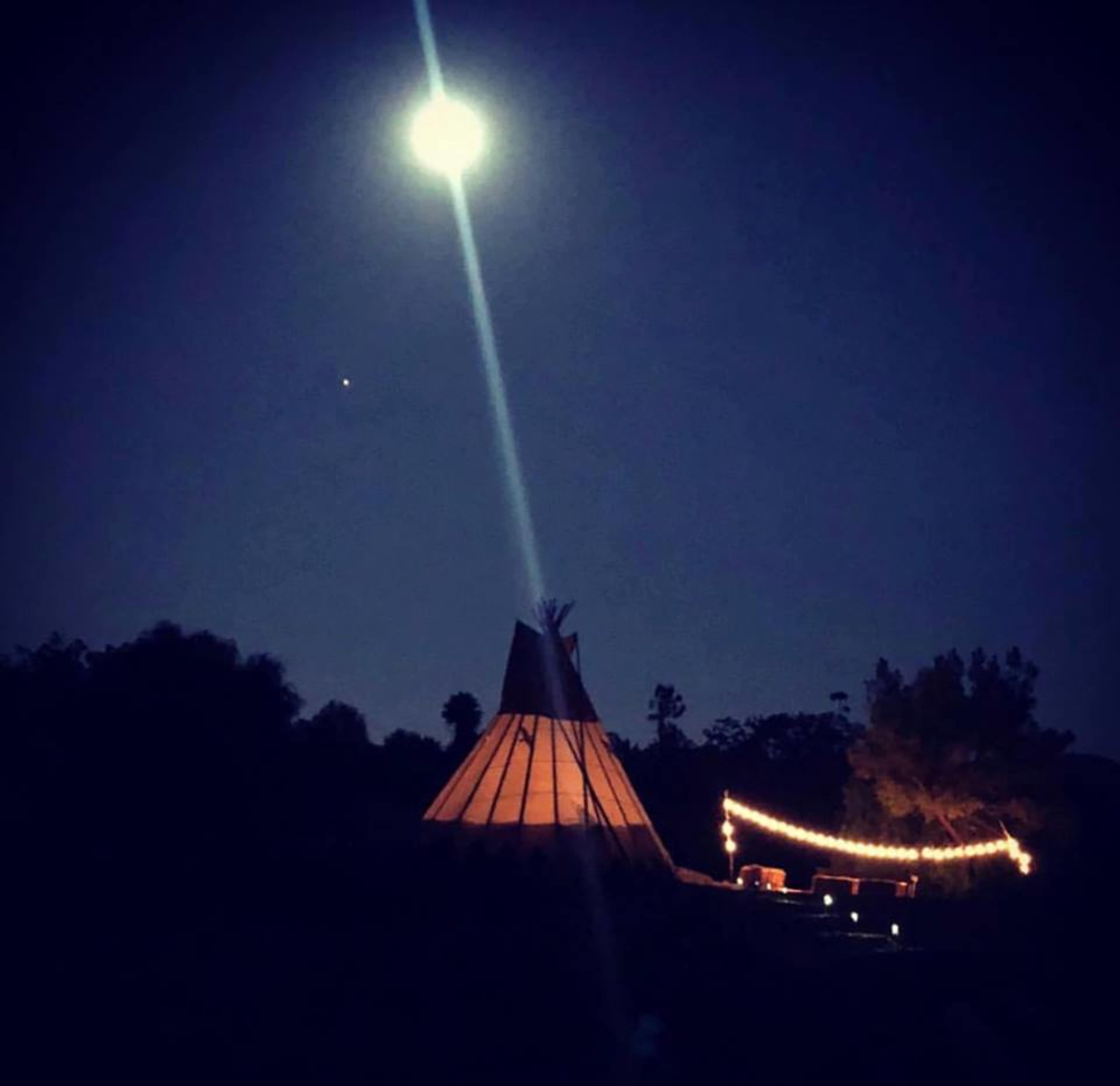 A lit teepee stands under a bright full moon, with string lights draped nearby against a dark sky.