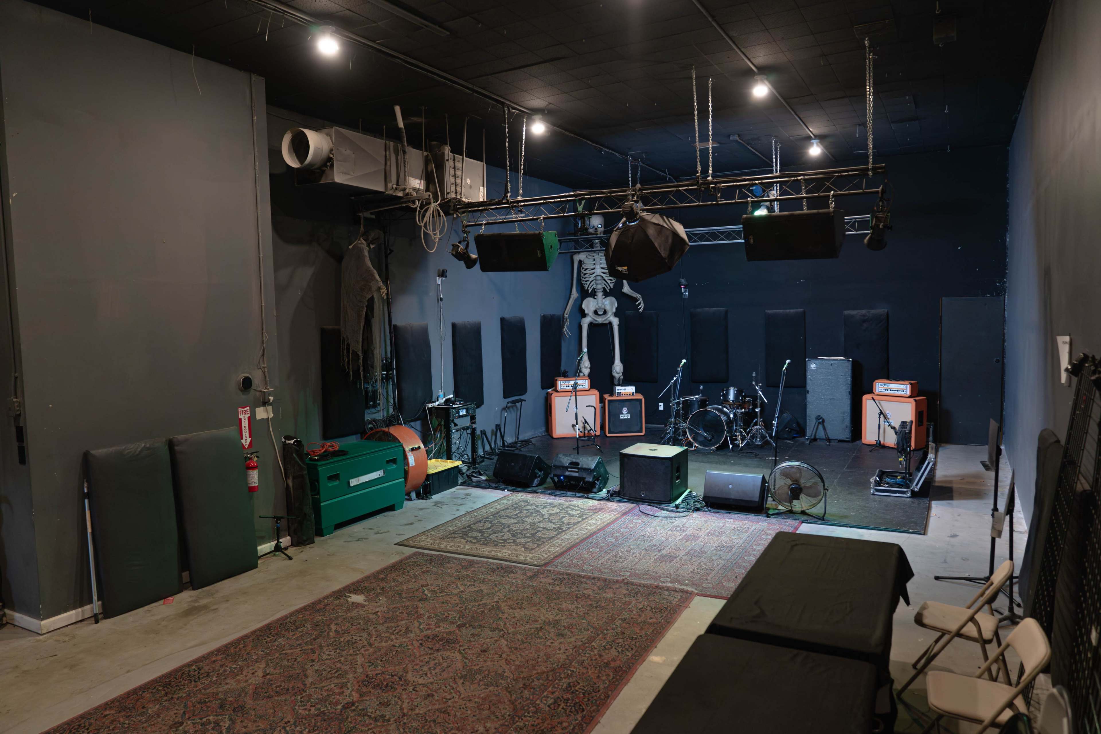 The image shows an interior view of a small music venue featuring a stage with musical instruments, amplifiers, and a decorative skeleton hanging in the background.