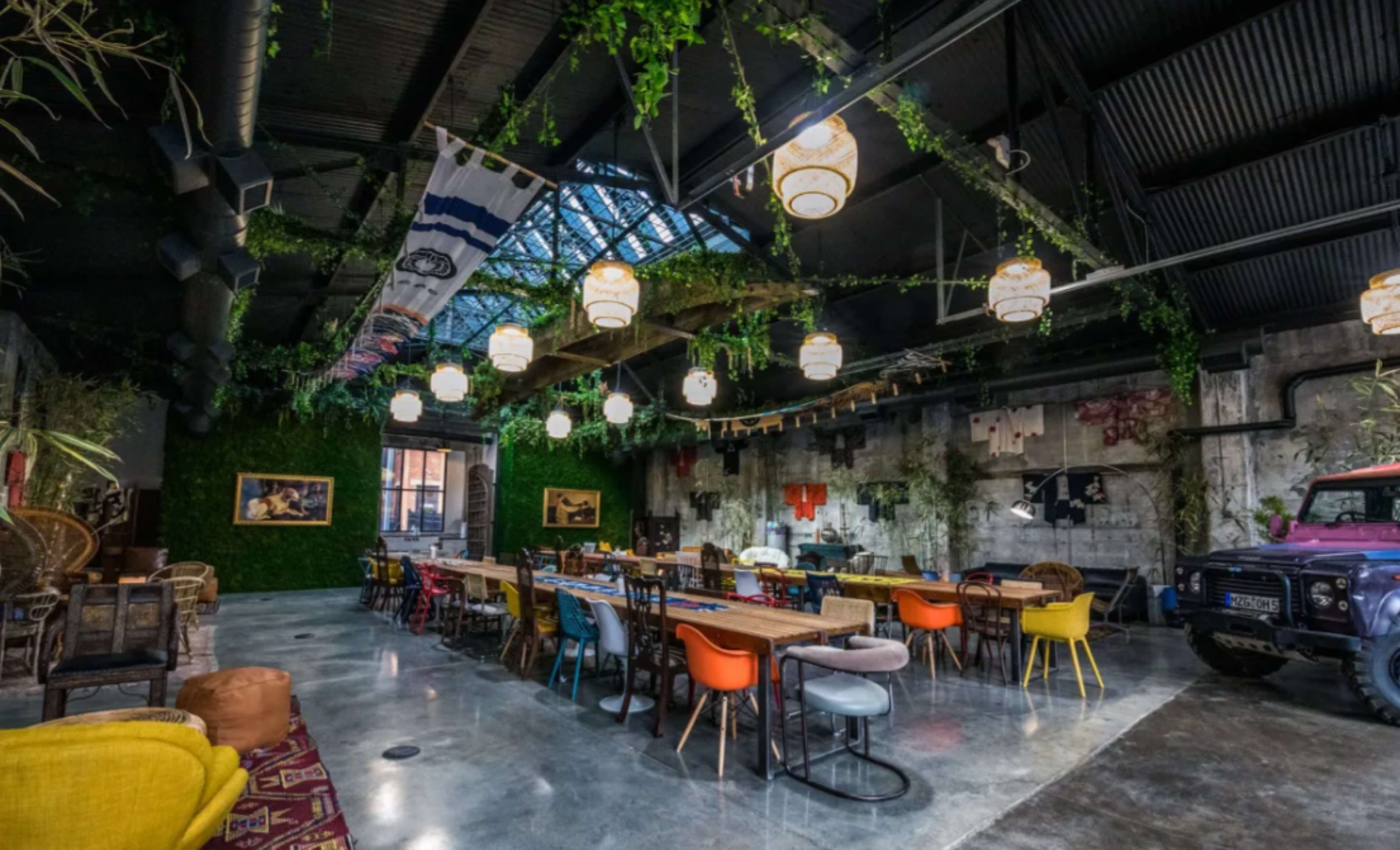 The image shows a spacious, modern café interior with a high ceiling, plants hanging from the beams, and a mix of colorful chairs around large wooden tables.