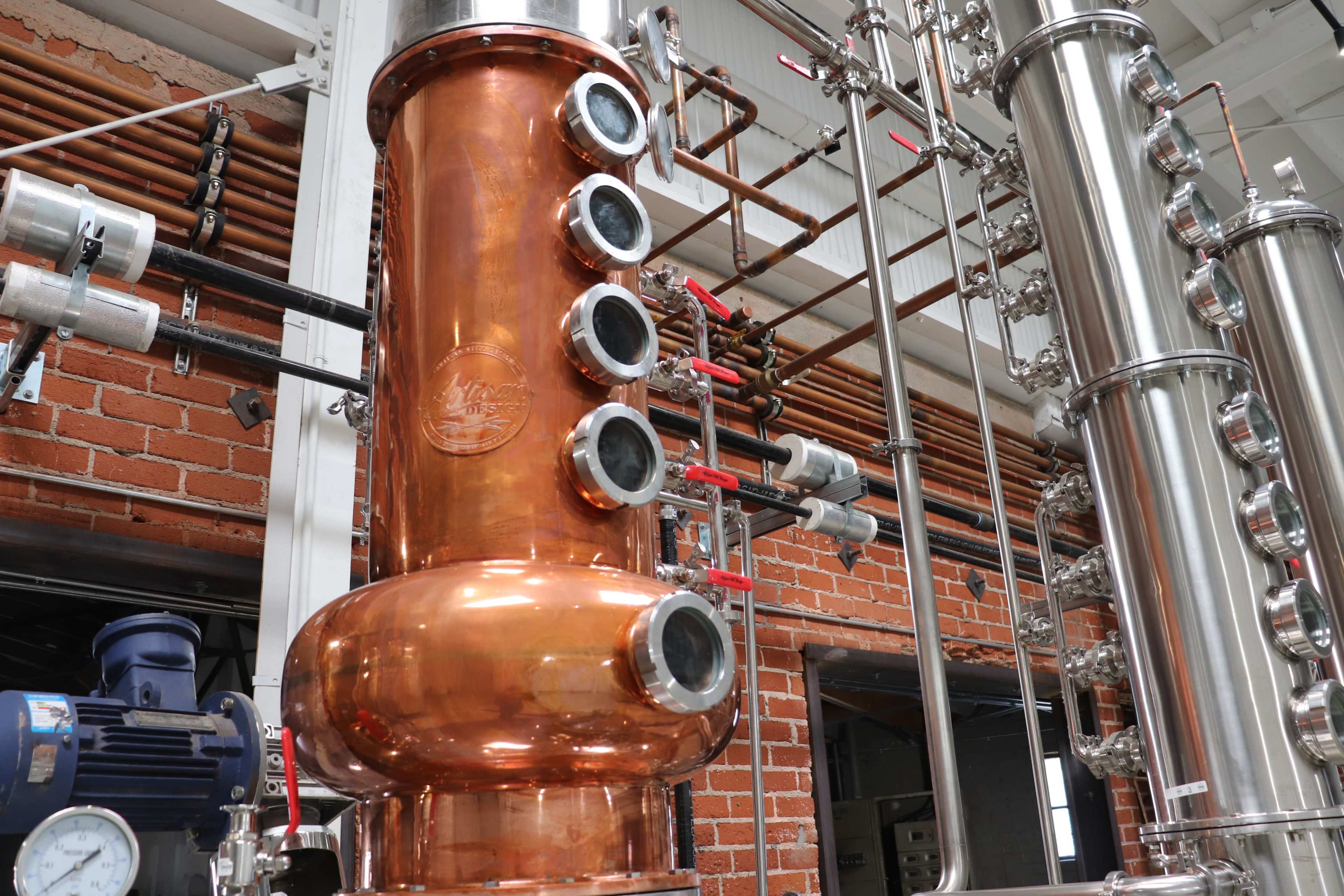 A copper distillation column with multiple viewing ports and connected piping in an industrial distillery setting.