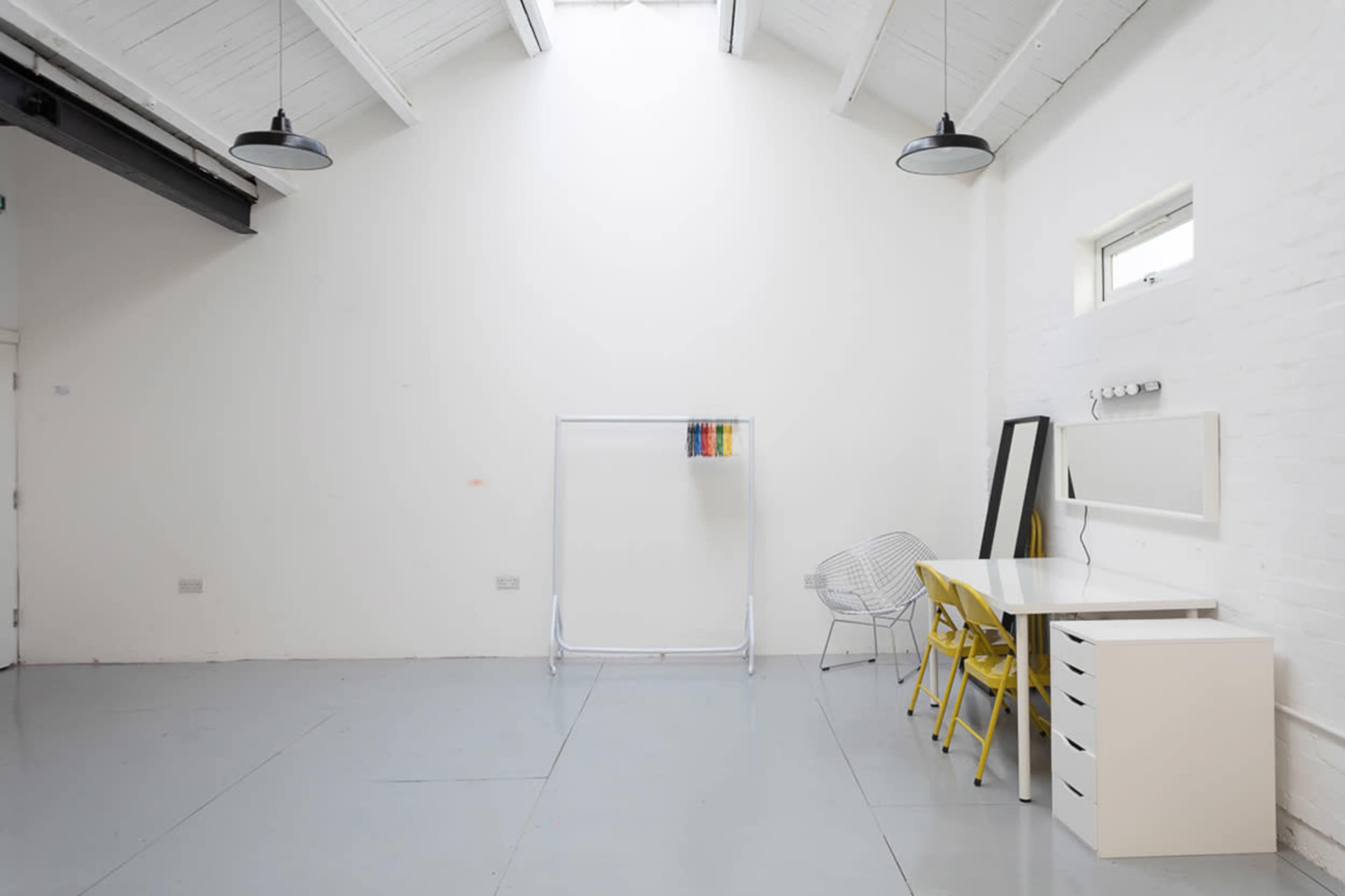 A minimalist interior space with white walls, a large window, a clothing rack, a mirror, a table with yellow chairs, and a light fixture.